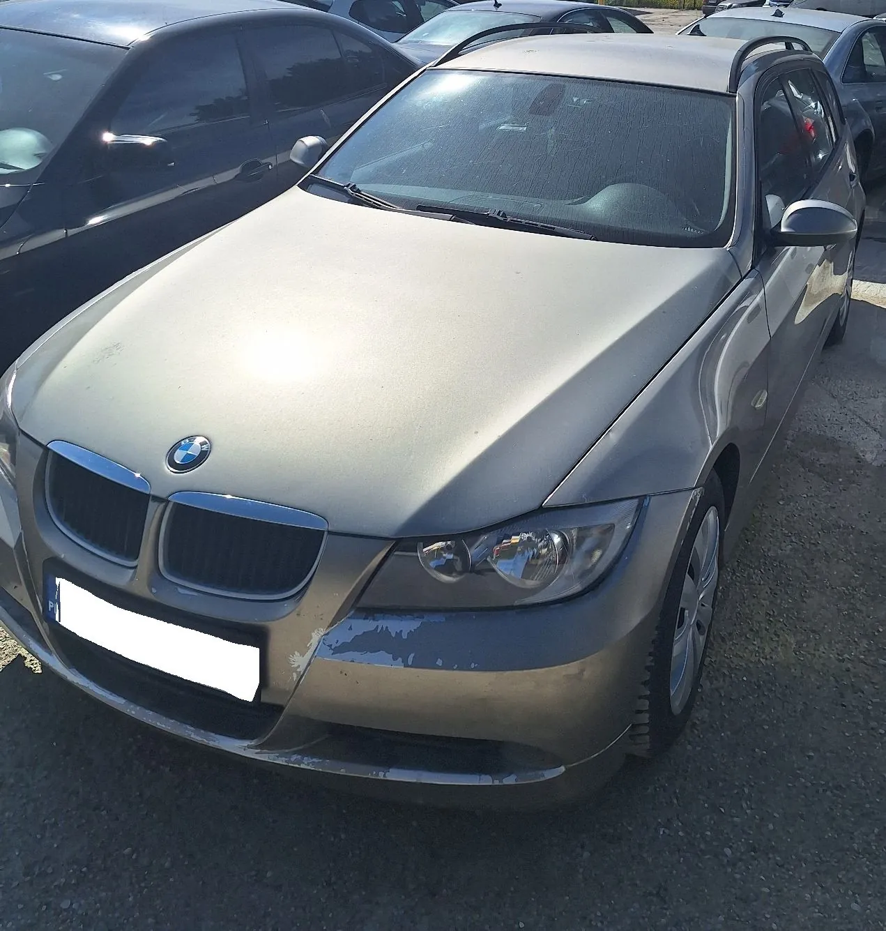 Srebrny samochód marki BMW zaparkowany na placu. Pojazd ma widoczne uszkodzenia lakieru na przednim zderzaku po lewej stronie. W tle widoczne są inne zaparkowane samochody.