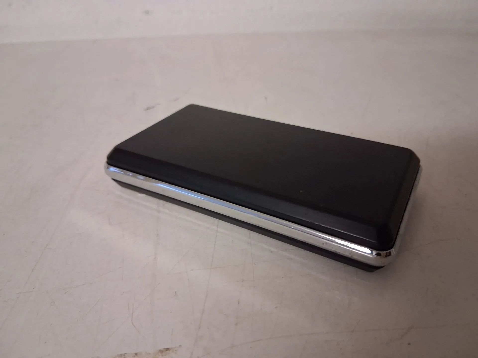 Czarny, prostokątny przedmiot o zaokrąglonych rogach, przypominający etui lub powerbank, z błyszczącą, srebrną ramką u dołu, leżący na jasnej, lekko porysowanej powierzchni.