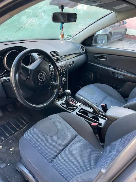 Wnętrze samochodu z widoczną kierownicą z logo Mercedes-Benz, szarymi siedzeniami w tkaninie, centralną konsolą z dźwignią zmiany biegów, przyciskami sterowania oraz lusterkiem bocznym. Przez przednią szybę widoczny jest inny samochód.