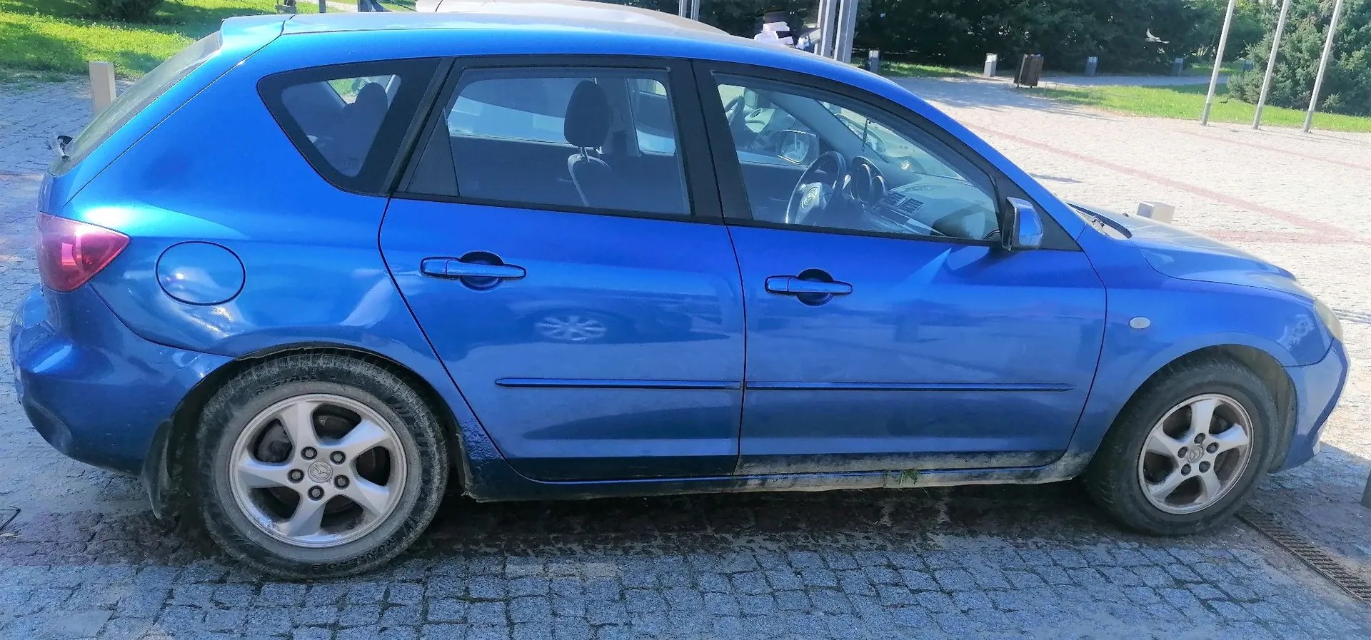 Niebieski samochód osobowy typu hatchback zaparkowany bokiem na kostce brukowej. Pojazd jest lekko zabrudzony w dolnych partiach nadwozia.