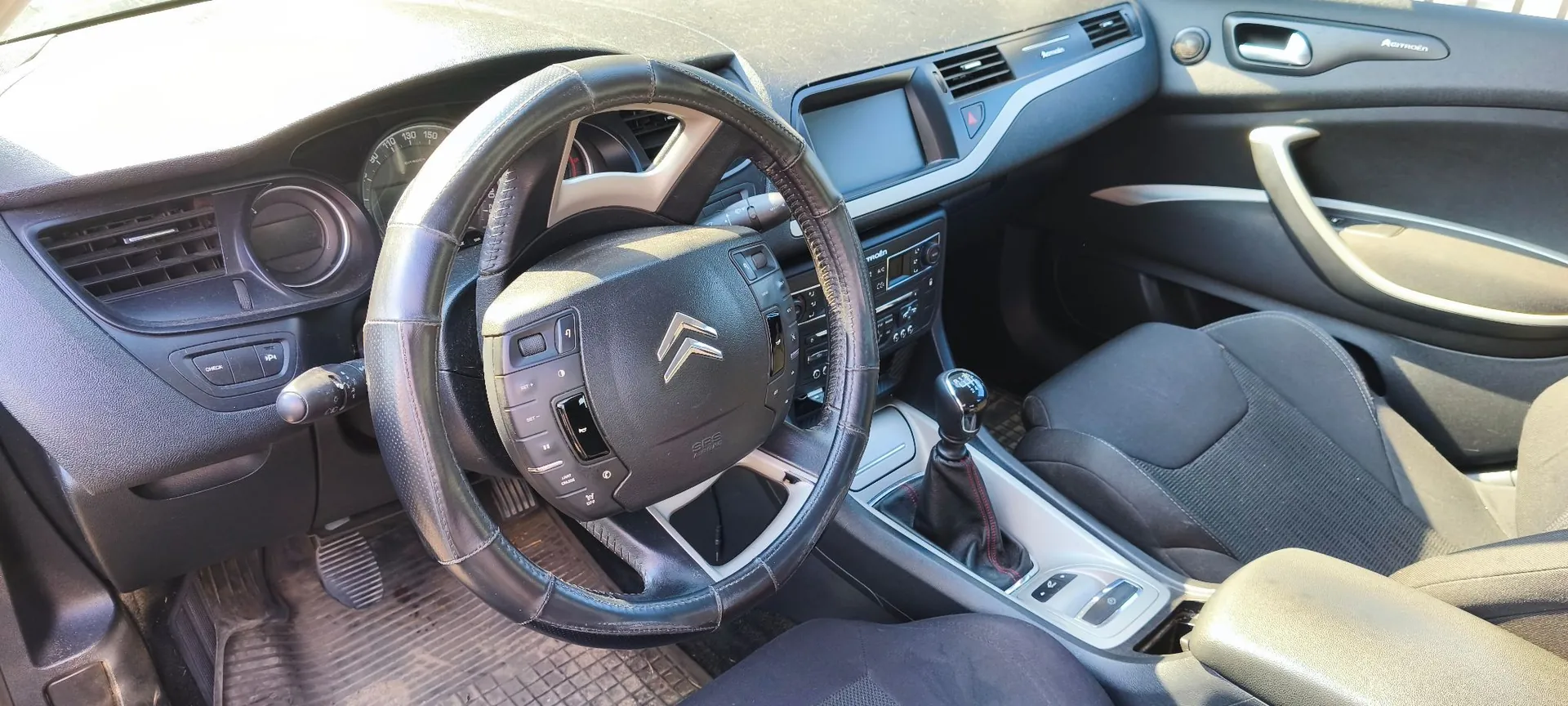 Wnętrze samochodu marki Citroën z perspektywy kierowcy. Widoczna jest czarna kierownica obszyta skórą z logo marki na środku, deska rozdzielcza z ekranem multimedialnym, zegary oraz czarne, materiałowe fotele. W tle widoczny jest fragment drzwi pasażera i konsola środkowa z lewarkiem zmiany biegów.