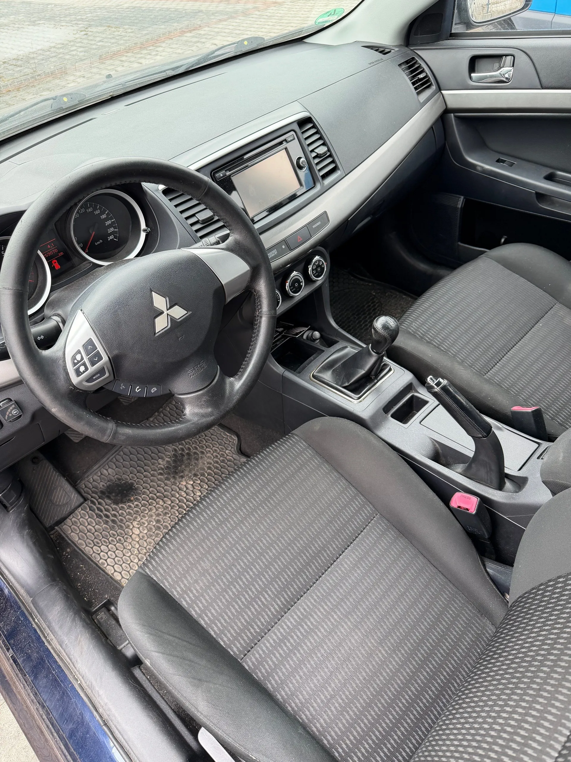 Wnętrze samochodu marki Mitsubishi z widocznym kierownicą z logo trzech rombów, czarnymi siedzeniami w paski, deska rozdzielcza z ekranem centralnym, wskaźnikami prędkości i obrotomierzem. Widoczne są również dźwignia zmiany biegów, pedały oraz otwarte drzwi kierowcy. Na podłodze znajdują się maty z wzorem. Przez przednią szybę widać brukowaną powierzchnię.