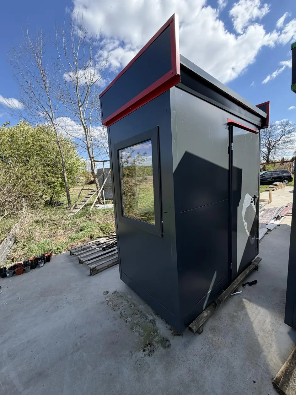 Ciemnoszara budka z czerwonymi akcentami na dachu, z oknem pośrodku. Budka stoi na betonowej powierzchni, w tle niebo z chmurami, drzewa bez liści, zielone krzewy, stosy drewnianych palet oraz pojazd. Na budce widoczne są ślady uszkodzeń i białe plamy.