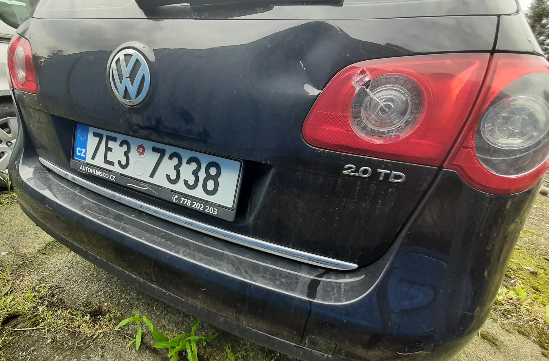 Tył czarnego samochodu marki Volkswagen z widocznym logo, tablicą rejestracyjną CZ 7E3 7338, napisem AUTOHLINSKO.CZ oraz numerem telefonu 778 202 203. Po prawej stronie widoczny napis 2.0 TD. Samochód stoi na podłożu z drobnej ziemi i trawy, z czerwonymi światłami tylnymi i lekkimi uszkodzeniami na karoserii.