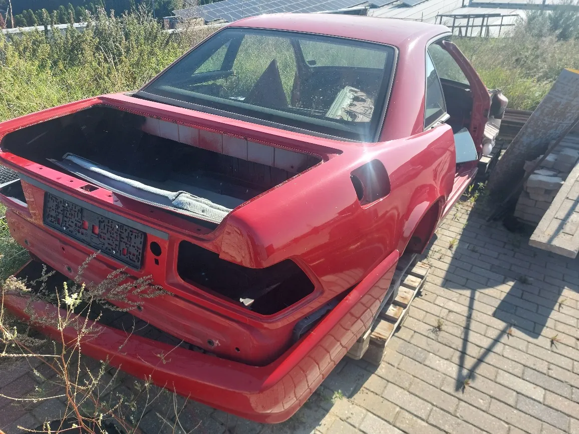 Tylna część czerwonego samochodu osobowego bez klapy bagażnika i tylnych lamp. Pojazd stoi na betonowej kostce brukowej obok trawy i krzewów. Wnętrze bagażnika jest puste, a w miejscu tablicy rejestracyjnej widoczny jest czarny plastikowy uchwyt.