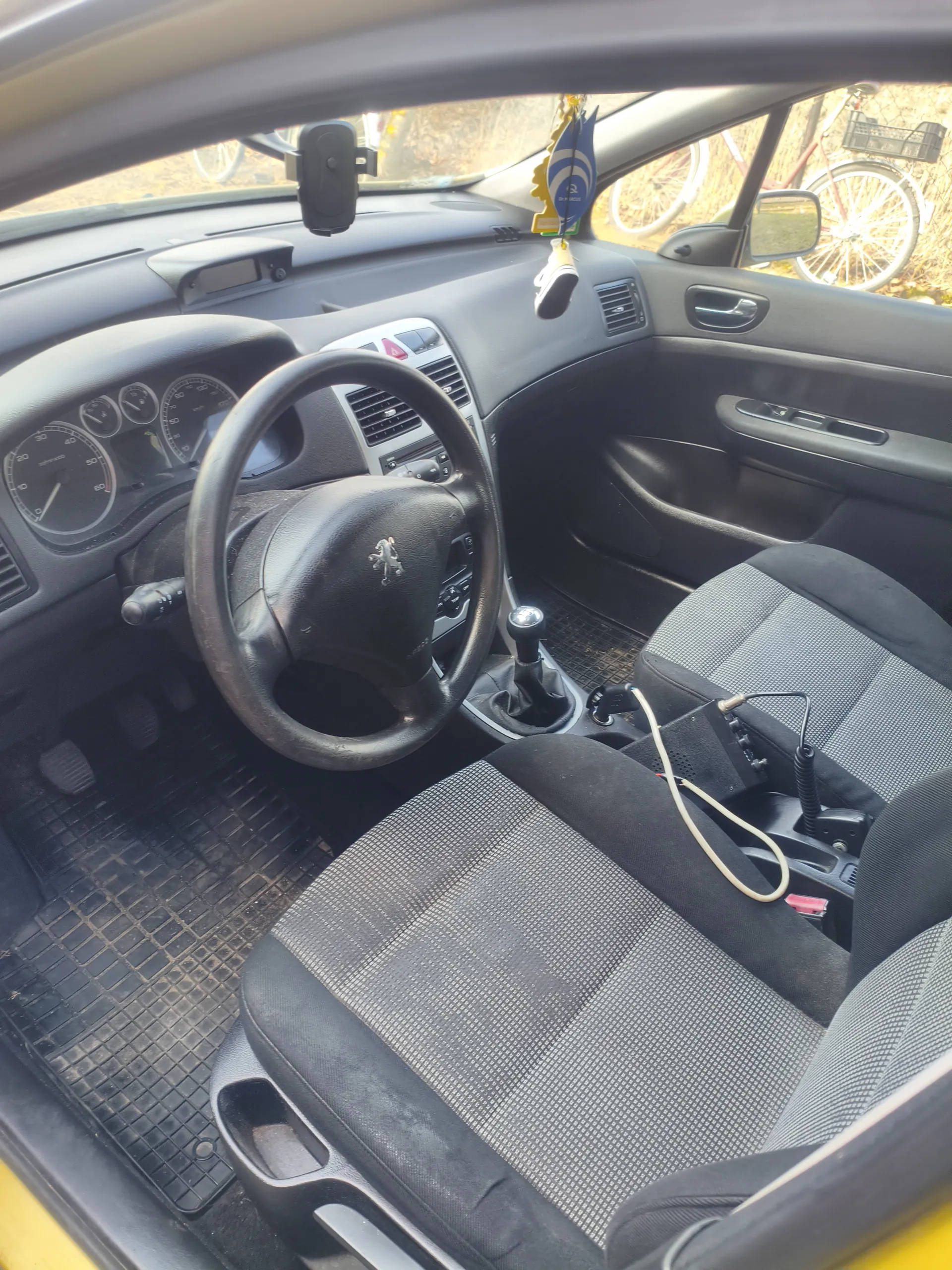 Wnętrze samochodu z widocznym kierownicą z logo, tablicą rozdzielczą z licznikami, dźwignią zmiany biegów i siedzeniami w czarno-szarym kolorze. Na siedzeniu pasażera znajduje się urządzenie z przewodami. Przez przednią szybę widoczne są rowery i ogrodzenie. Wnętrze posiada czarne wykończenie, a na desce rozdzielczej umieszczono wiszące ozdoby.