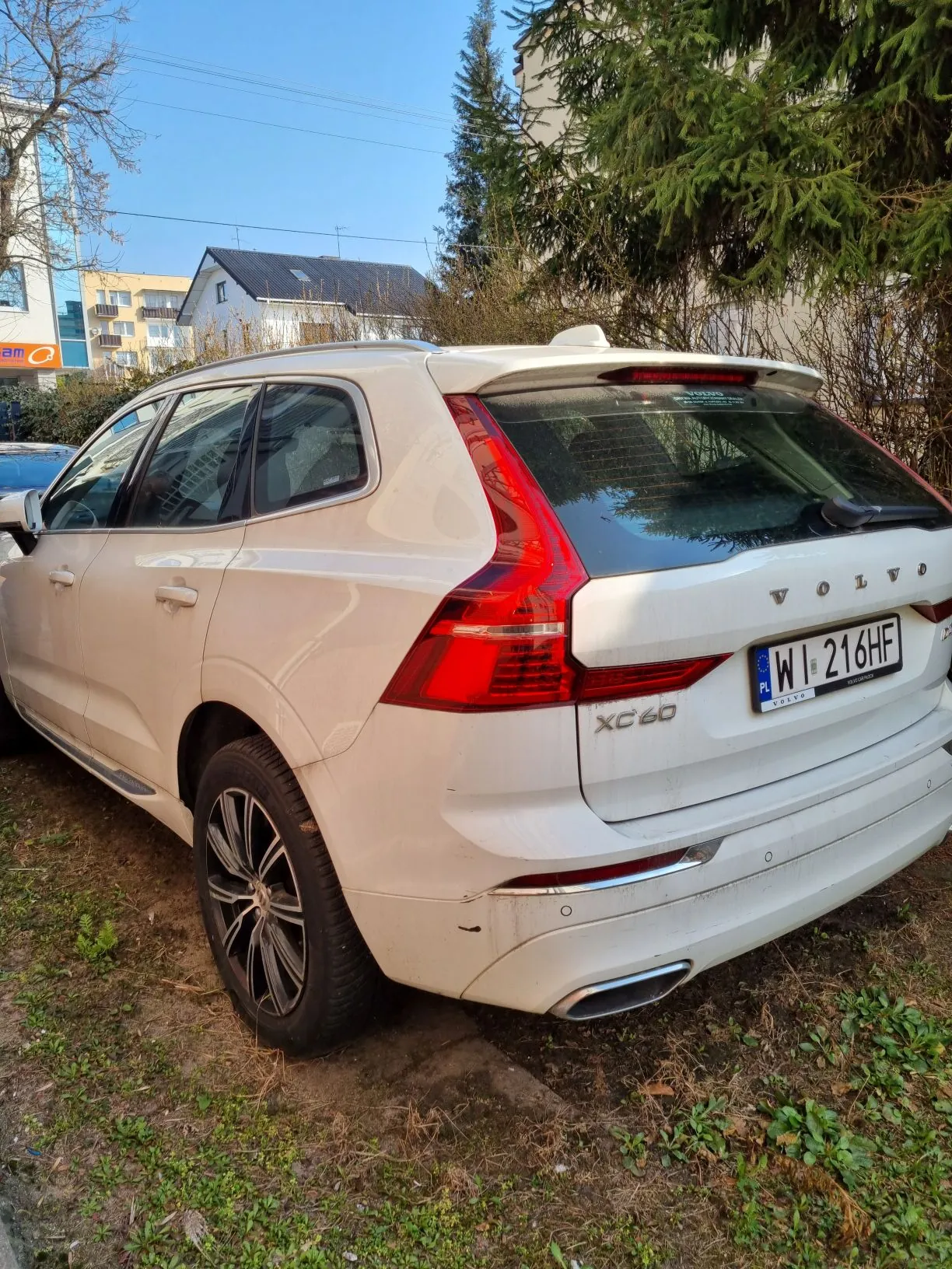 Biały samochód Volvo XC60 zaparkowany na trawie obok drzew i budynków. Widoczna tablica rejestracyjna WI 216HF. Tło z jasnym niebem i budynkami mieszkalnymi.