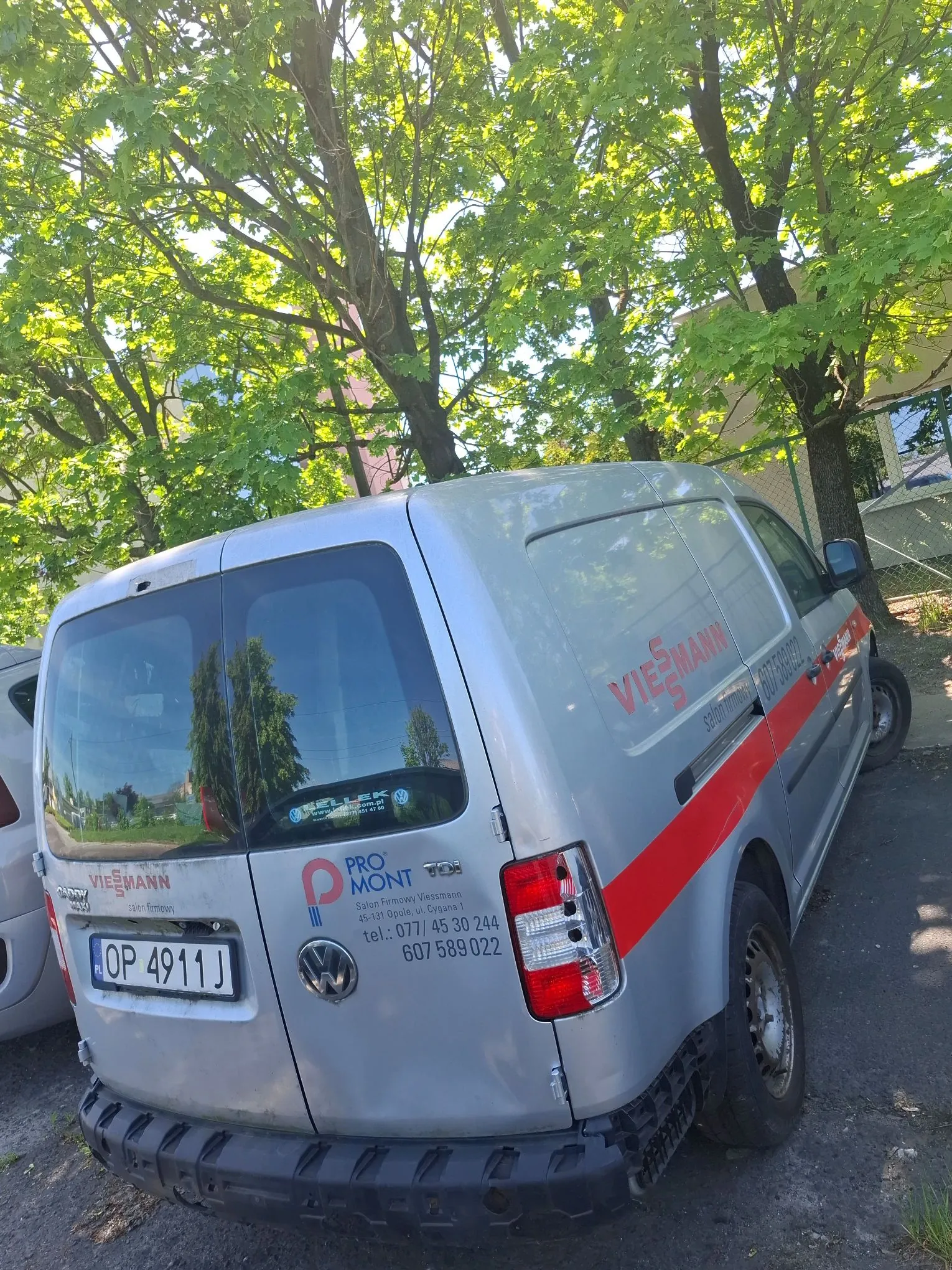 Srebrny samochód dostawczy marki Volkswagen zaparkowany pod drzewami z zielonymi liśćmi. Na boku i tylnych drzwiach pojazdu znajdują się napisy: 'VIESSMANN', 'PROMONT', 'Salon Firmowy Viessmann', adres '45-120 Bochnia ul. Sypna 1' oraz numery telefonów 'tel.: 077/ 45 30 244' i '607 589 022'. Tablica rejestracyjna samochodu to OP 4911J.