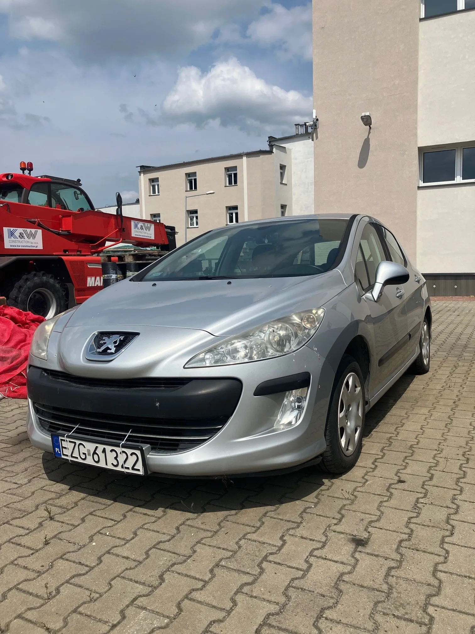 Srebrny samochód osobowy marki Peugeot zaparkowany na kostce brukowej. W tle widoczny jest czerwony pojazd specjalistyczny z logo 'K&W' oraz jasny, wielopiętrowy budynek mieszkalny pod zachmurzonym niebem. Samochód posiada tablicę rejestracyjną o numerze EZG 61322.
