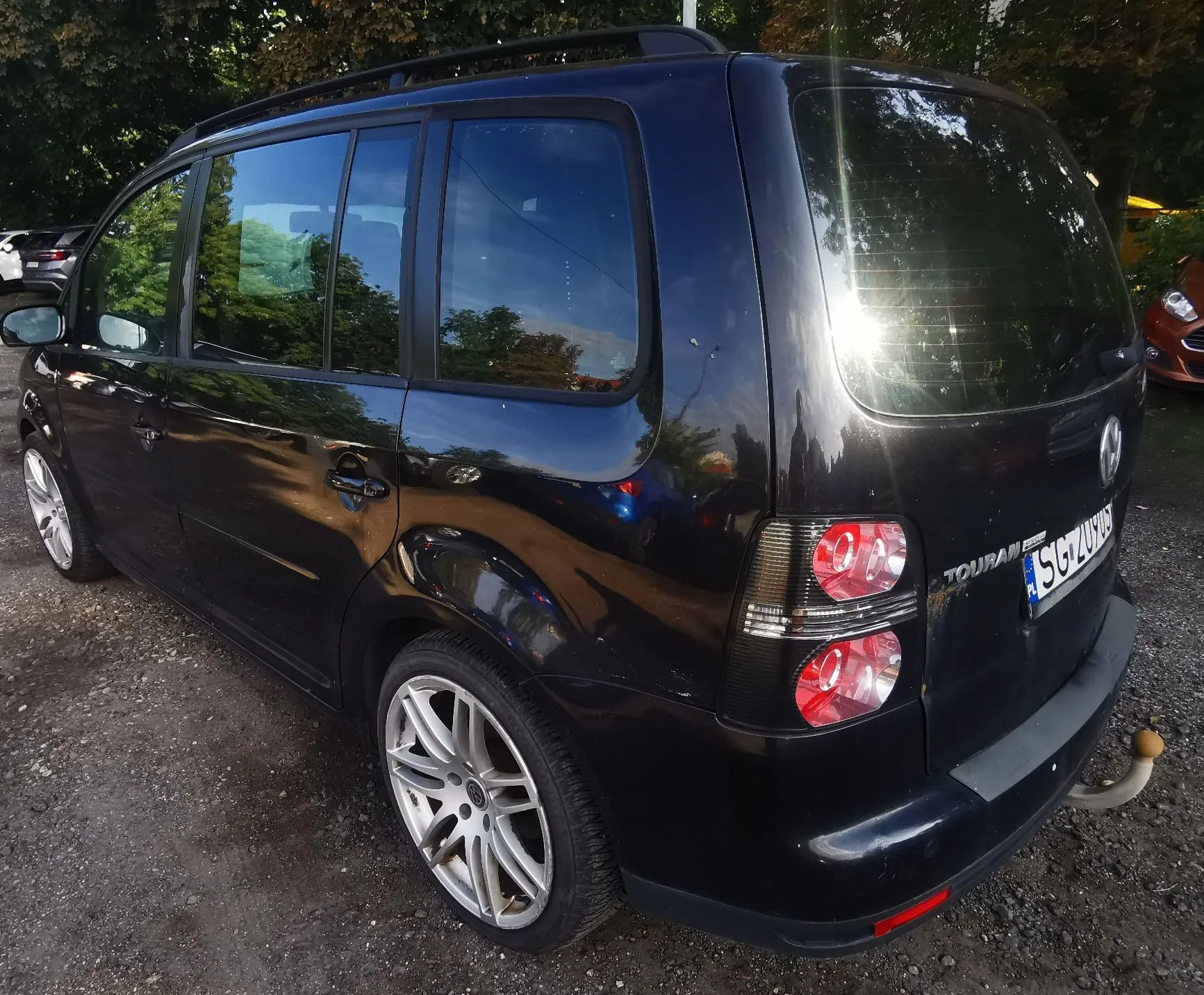 Czarny samochód marki Volkswagen Touran zaparkowany na żwirowym podłożu. Widok z tyłu i z boku pojazdu. Samochód posiada srebrne felgi aluminiowe, relingi dachowe oraz hak holowniczy. Na klapie bagażnika widnieje napis TOURAN oraz tablica rejestracyjna SGZ 209... (częściowo zasłonięta).