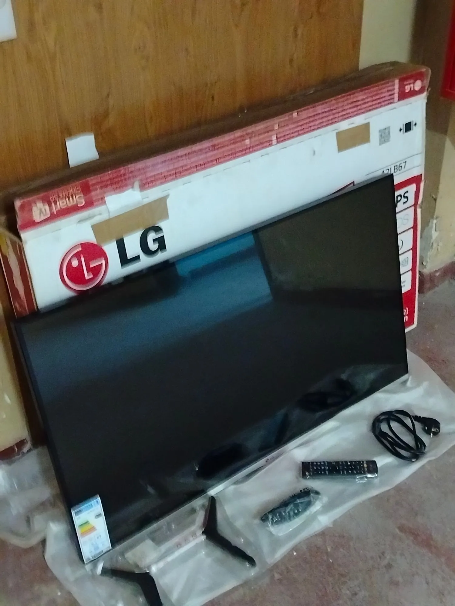 Czarny telewizor LG oparty o swoje oryginalne, biało-czerwone opakowanie kartonowe. Przed telewizorem na białej folii ochronnej leży pilot do sterowania oraz zestaw śrub w plastikowym woreczku. Na kartonie widoczne jest logo LG oraz napis 'Smart'.