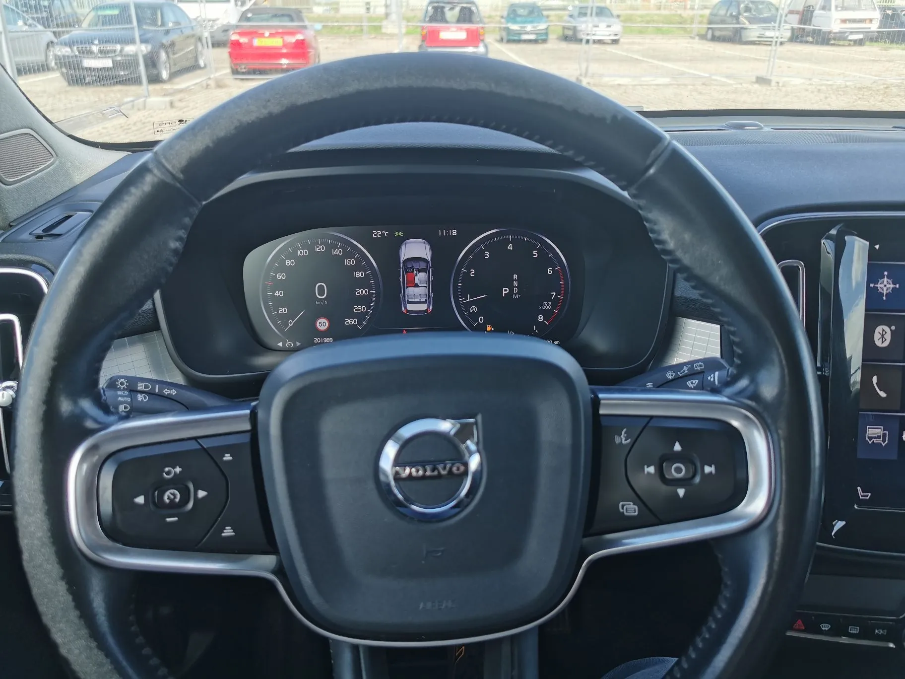 Widok z perspektywy kierowcy wnętrza samochodu marki Volvo. Na pierwszym planie znajduje się czarna, skórzana kierownica z logo Volvo na środku oraz przyciskami sterującymi po obu stronach. W tle widoczny jest cyfrowy zestaw wskaźników z prędkościomierzem, obrotomierzem oraz graficznym przedstawieniem pojazdu. Przez przednią szybę widać parking z zaparkowanymi samochodami w różnych kolorach oraz ogrodzenie z siatki.