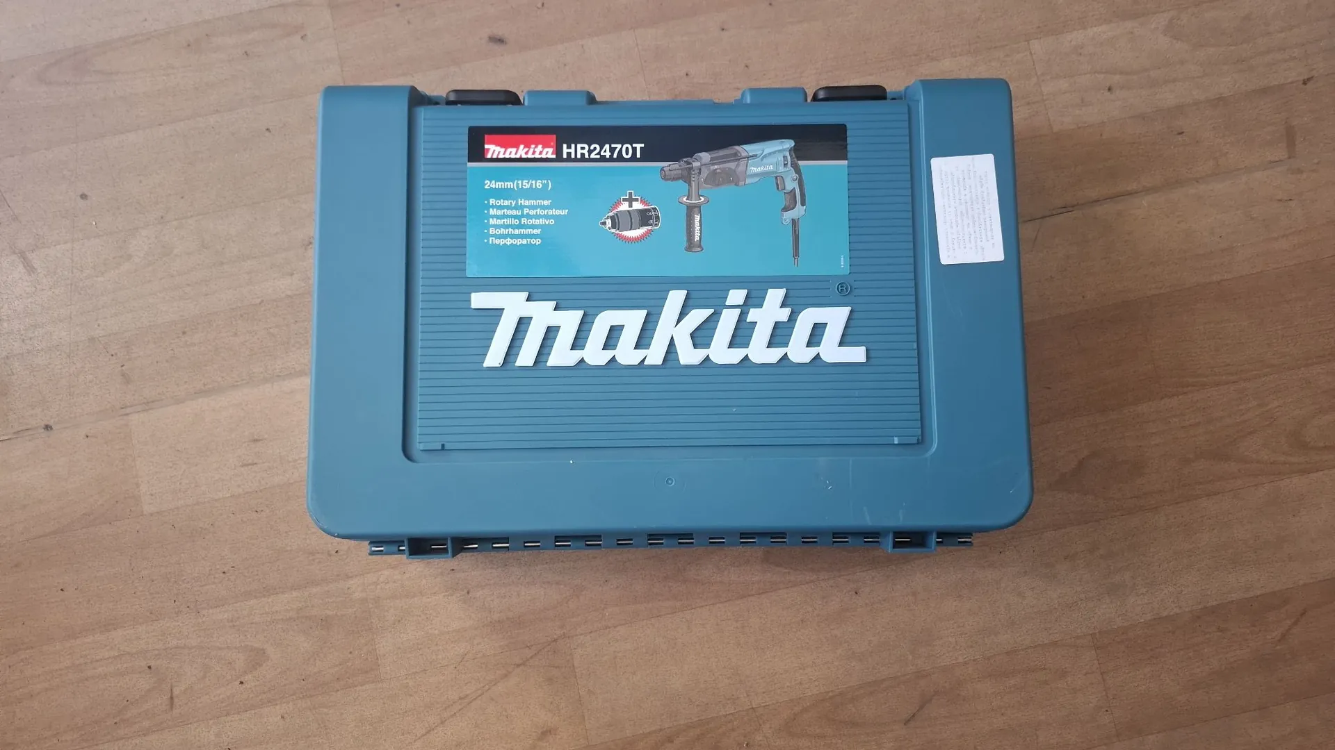 Niebieska plastikowa walizka narzędziowa marki Makita leżąca na drewnianej podłodze. Na pokrywie znajduje się duże białe logo 'Makita' oraz grafika przedstawiająca młotowiertarkę z napisem 'HR2470T' i specyfikacją techniczną, w tym '24mm(15/16") - Rotary Hammer'. Tekst i logo są zorientowane do góry nogami.
