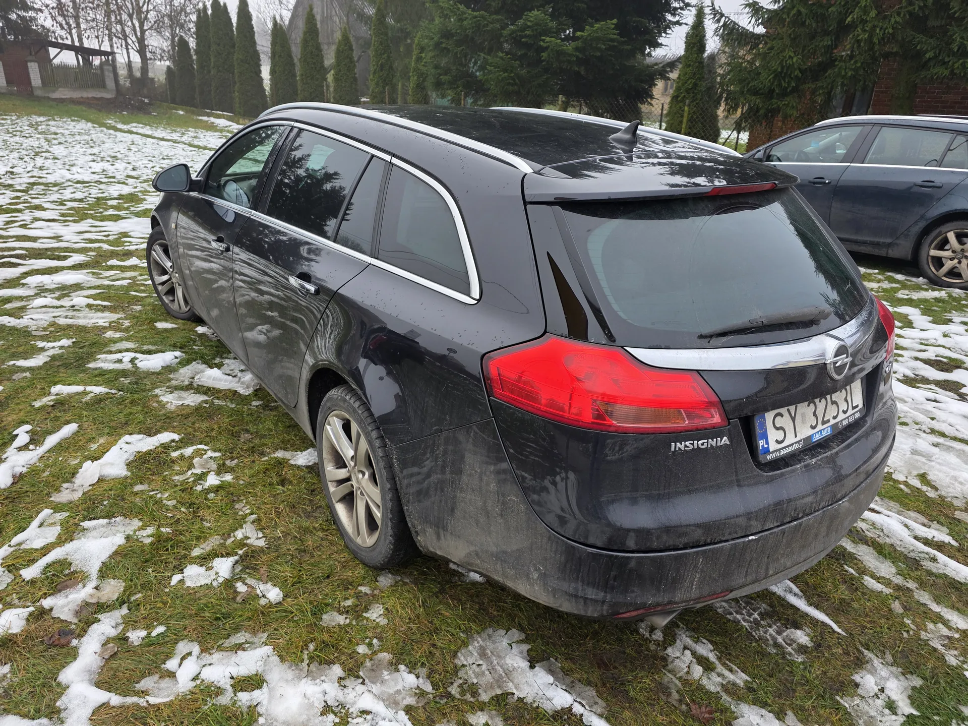 Czarny samochód typu kombi marki Opel Insignia zaparkowany na trawie z resztkami śniegu. Widoczny tył pojazdu z czerwonymi światłami, tablicą rejestracyjną SY 3253L oraz napisem INSIGNIA. W tle drugi ciemny samochód, drzewa iglaste, domy i ogrodzenie. Na tablicy rejestracyjnej widoczne oznaczenie PL oraz adres www.aaaauto.pl.