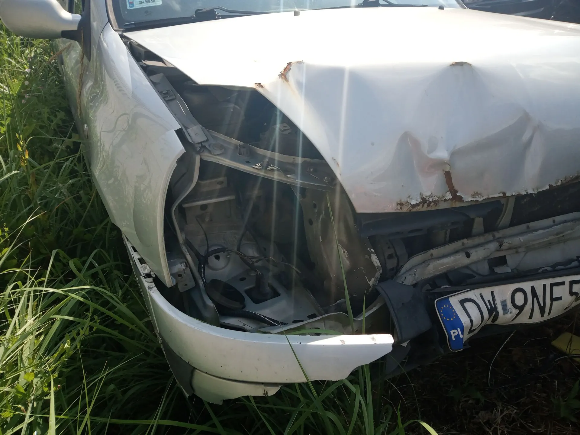 Zdjęcie przodu białego samochodu osobowego z poważnymi uszkodzeniami karoserii. Przednia maska jest wgnieciona i zagięta, brakuje prawego reflektora, a zderzak jest częściowo oderwany i pęknięty, odsłaniając elementy konstrukcyjne pojazdu. Samochód stoi na trawie. Widoczny jest fragment tablicy rejestracyjnej z oznaczeniem PL oraz literami DW 9NF.