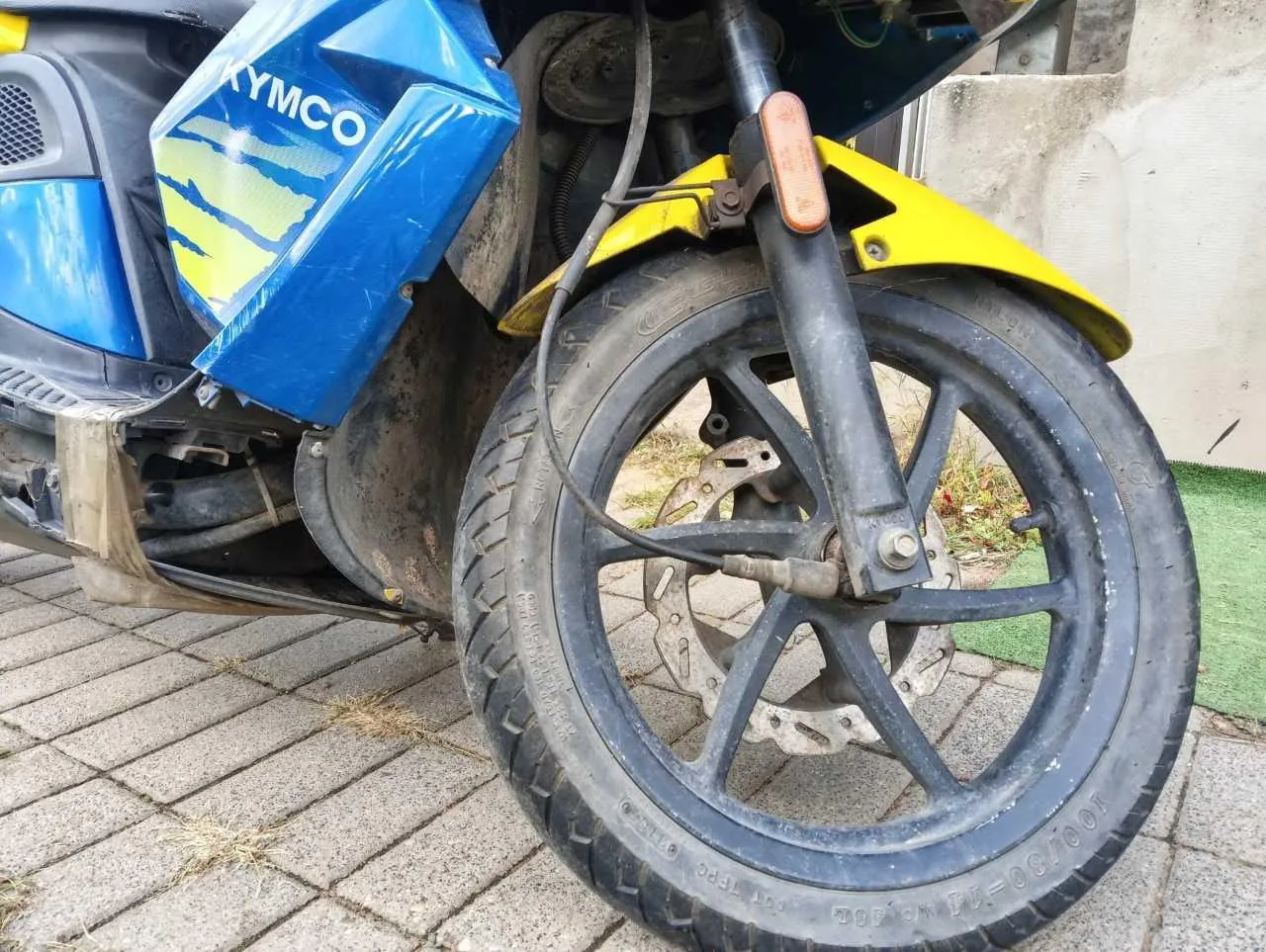 Zbliżenie na przednie koło niebiesko-żółtego skutera marki KYMCO. Widoczna jest czarna felga z oponą, tarcza hamulcowa oraz przedni amortyzator. Nad kołem znajduje się żółty błotnik i pomarańczowy kierunkowskaz. Skuter stoi na szarej kostce brukowej.