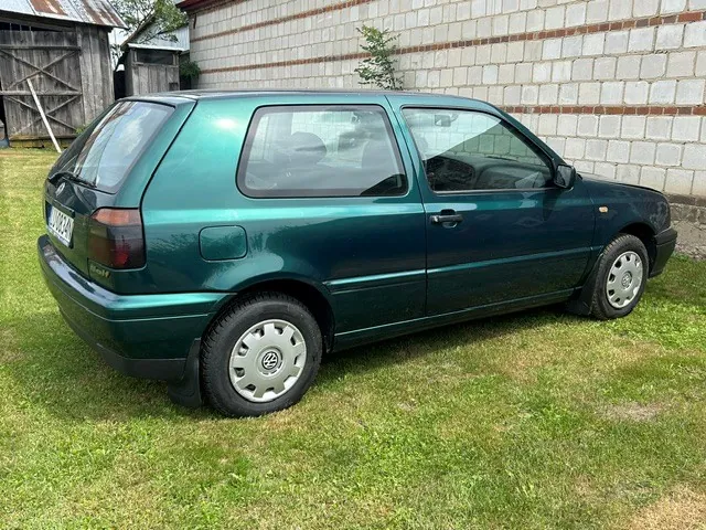 Zielony samochód marki Volkswagen Golf zaparkowany na trawie na tle białego muru z cegieł i drewnianego budynku.