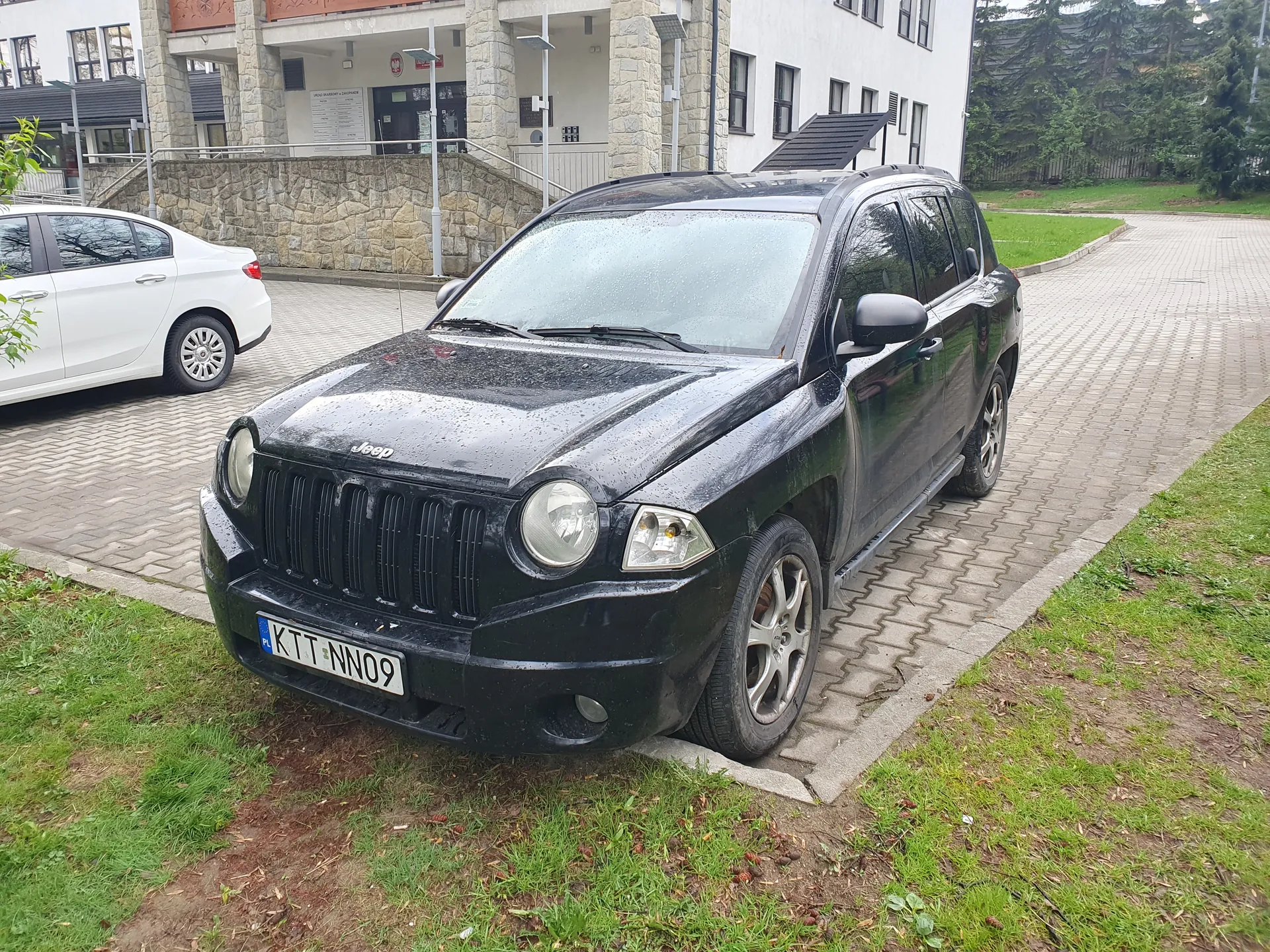 Czarny samochód Jeep z tablicą rejestracyjną KTT NN09 zaparkowany na brukowanym placu. Po lewej stronie widoczny biały samochód. W tle budynek z kamiennej cegły i betonu z wejściem oraz tablicą informacyjną. Na przedniej szybie i karoserii Jeepa krople deszczu. Teren wokół częściowo zielony, z trawą i brukiem.
