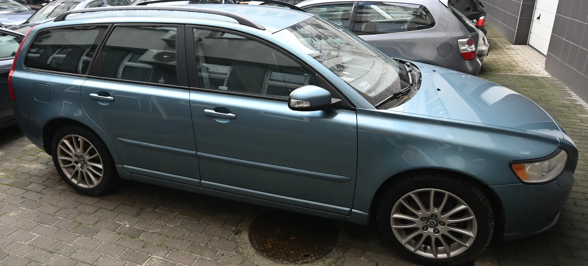 Jasnoniebieski samochód typu kombi Volvo zaparkowany na brukowanej powierzchni. Widoczne dachowe pręty, felgi aluminiowe, przyciemniane szyby. W tle inne pojazdy, budynek z bramą garażową oraz pokrywa kanału ściekowego na nawierzchni.