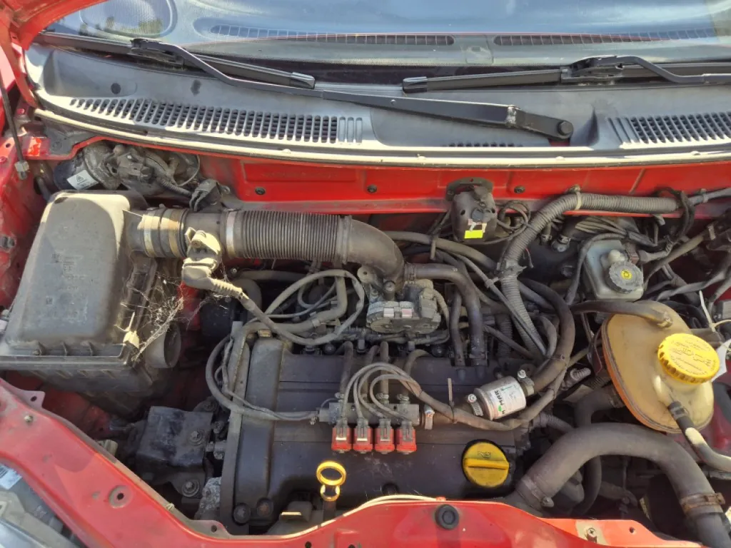 Widok z góry na komorę silnika w czerwonym samochodzie. Widoczne są elementy mechaniczne: czarna obudowa filtra powietrza po lewej stronie, rury dolotowe, przewody, akumulator oraz zbiornik z żółtym korkiem po prawej stronie. Na górze znajduje się przednia szyba z wycieraczkami.
