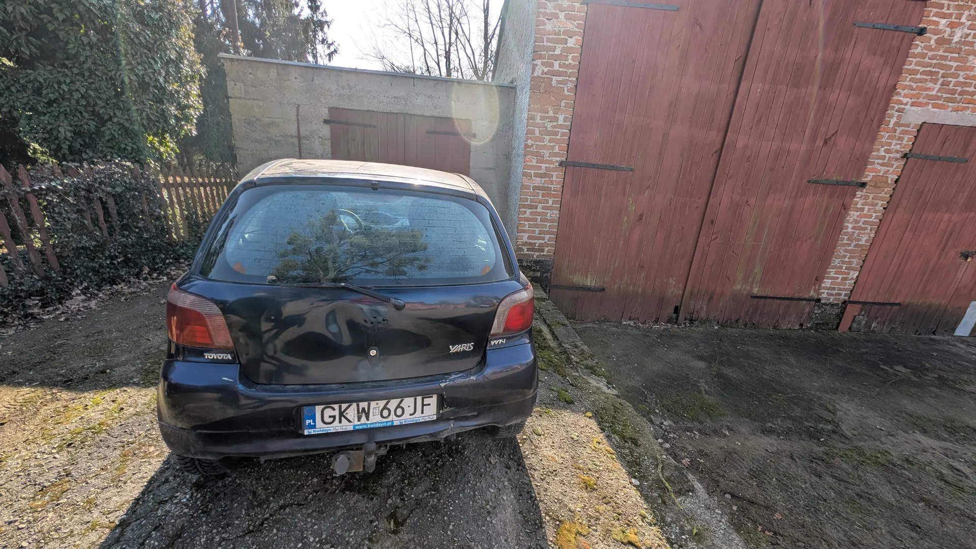 Czarny samochód Toyota Yaris z tablicą rejestracyjną PL GKW 66 JF zaparkowany tyłem na podwórku. W tle ceglany budynek z czerwonymi drewnianymi drzwiami garażowymi, betonowa ściana, ogrodzenie z drewnianych słupków oraz drzewa. Podłoga składa się z żwiru i asfaltu, na którym widoczne są plamy mchu.