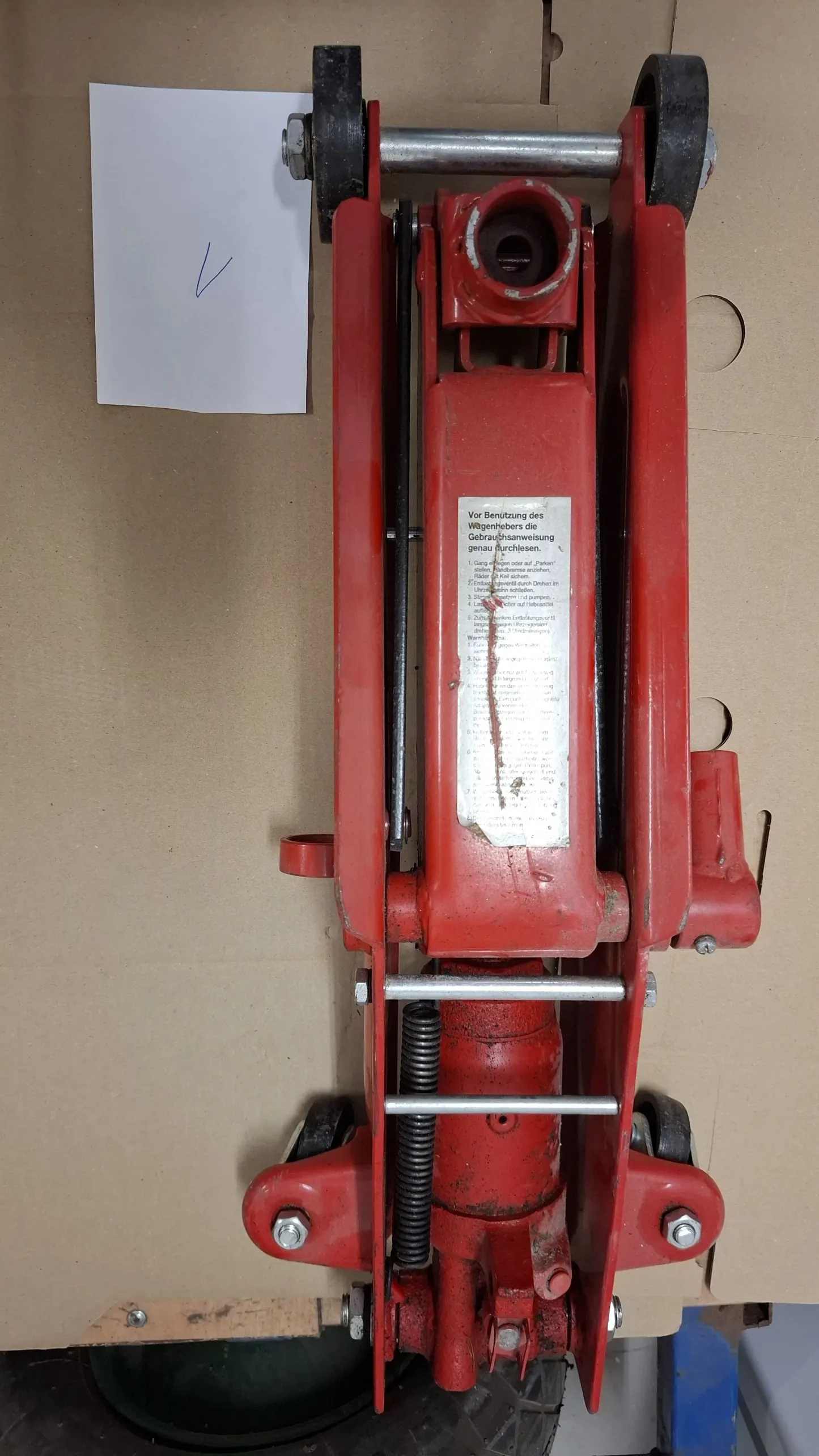 Czerwony podnośnik hydrauliczny do samochodów leżący na brązowym kartonie. Urządzenie jest ułożone pionowo w kadrze, z kółkami skierowanymi w górę i w dół. Na obudowie znajduje się biała tabliczka znamionowa z tekstem w języku niemieckim. Obok podnośnika leży biała kartka papieru z napisem '1'.