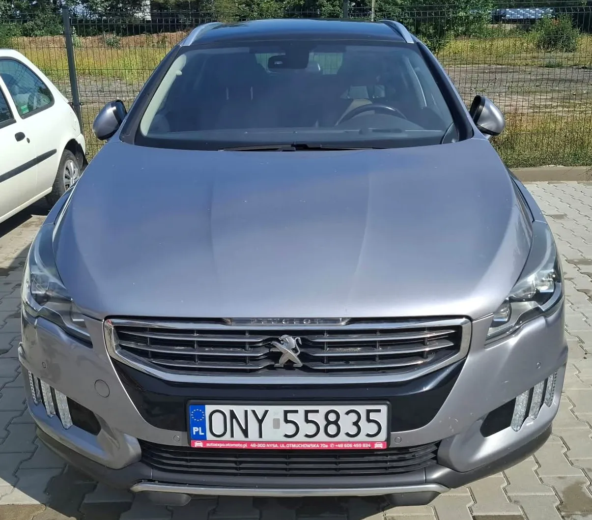Srebrny samochód marki Peugeot widziany od przodu, zaparkowany na kostce brukowej. Pojazd posiada tablicę rejestracyjną o numerze ONY 55835 z oznaczeniem PL. W tle widoczny jest biały samochód oraz zielony płot i roślinność.