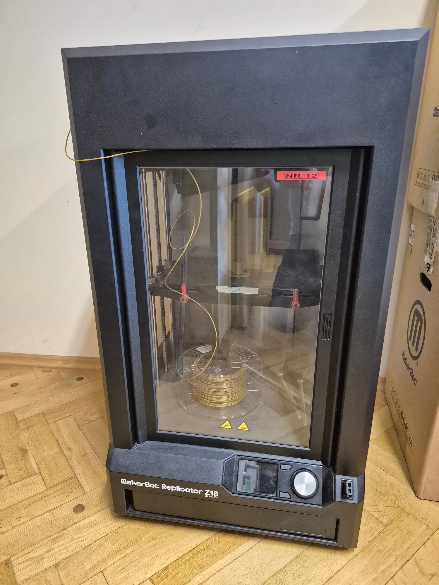 Czarna drukarka 3D marki MakerBot Replicator Z18 w zamkniętej obudowie z przeszklonym frontem. Wewnątrz urządzenia widoczna jest szpula z żółtym filamentem oraz mechanizmy drukujące. Na dolnej ramie znajduje się napis 'MakerBot Replicator Z18'. W prawym górnym rogu szyby przyklejona jest czerwona etykieta z napisem 'NR-12'. Urządzenie stoi na drewnianej podłodze.
