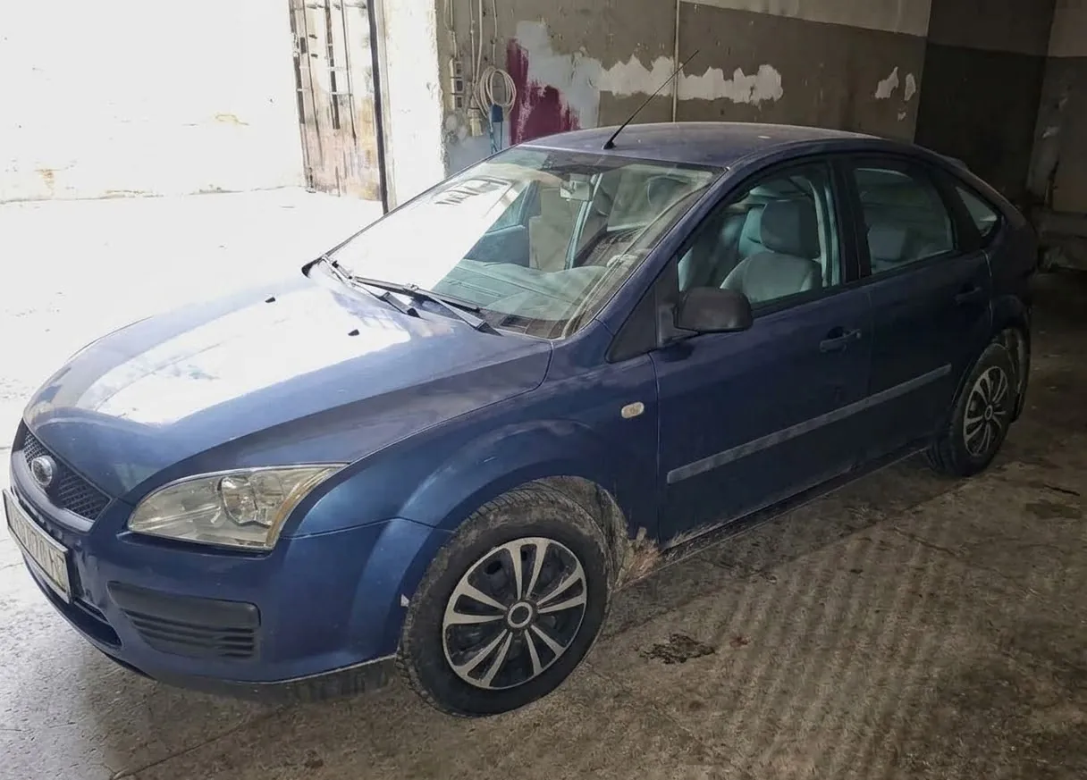 Niebieski samochód osobowy marki Ford Focus zaparkowany w garażu lub warsztacie. Pojazd jest widoczny z przodu i z boku, ma srebrne felgi aluminiowe i jest lekko zabrudzony. W tle widać betonową ścianę i otwarcie na zewnątrz.