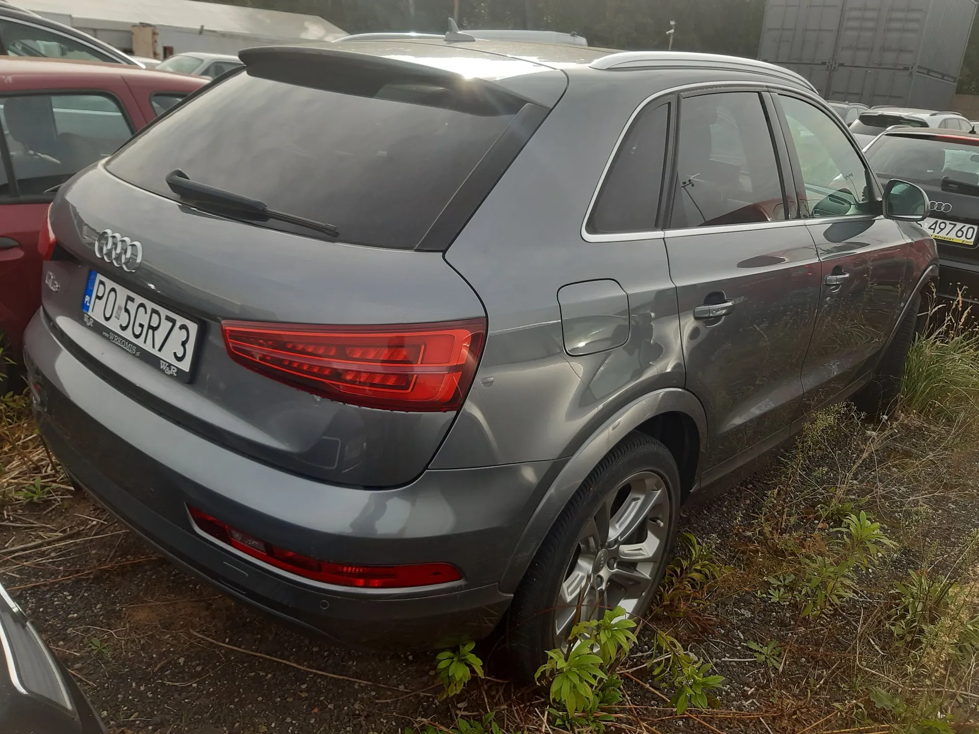 Tylny widok szarego samochodu marki Audi z tablicą rejestracyjną PO 5GR73. Pojazd zaparkowany jest na nieutwardzonym terenie z trawą i suchymi roślinami. W tle widoczne są inne samochody.