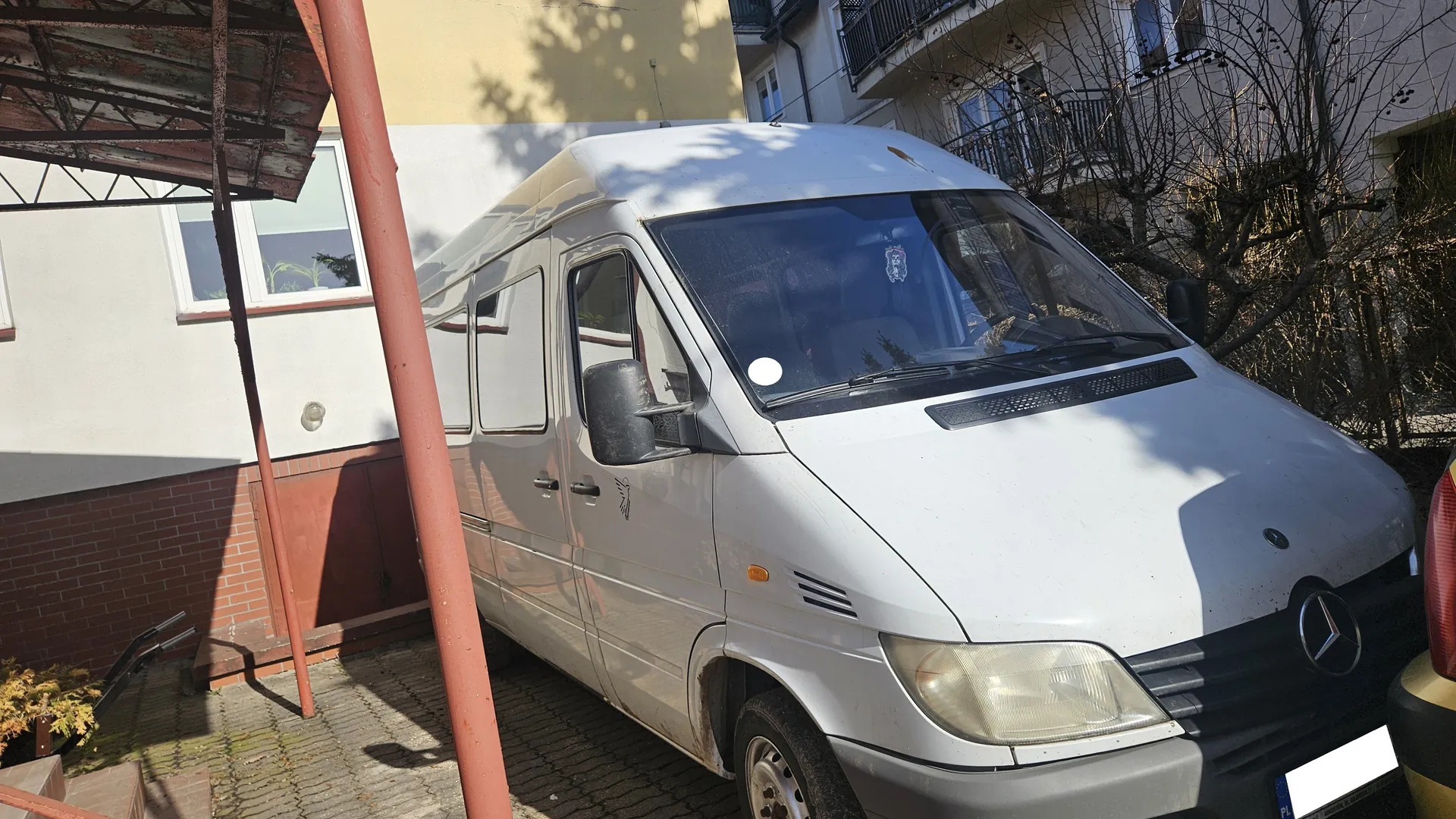 Srebrny samochód dostawczy marki Mercedes-Benz zaparkowany na brukowanym podwórku. Po lewej stronie budynek z białą i ceglaną ścianą, oknami oraz metalowym daszkiem. W tle widoczne są drzewa bez liści i fragmenty innych budynków. Na przedniej szybie samochodu naklejka, a na tablicy rejestracyjnej widoczny fragment oznaczenia PL. Oświetlenie słoneczne rzuca cienie na pojazd i otoczenie.
