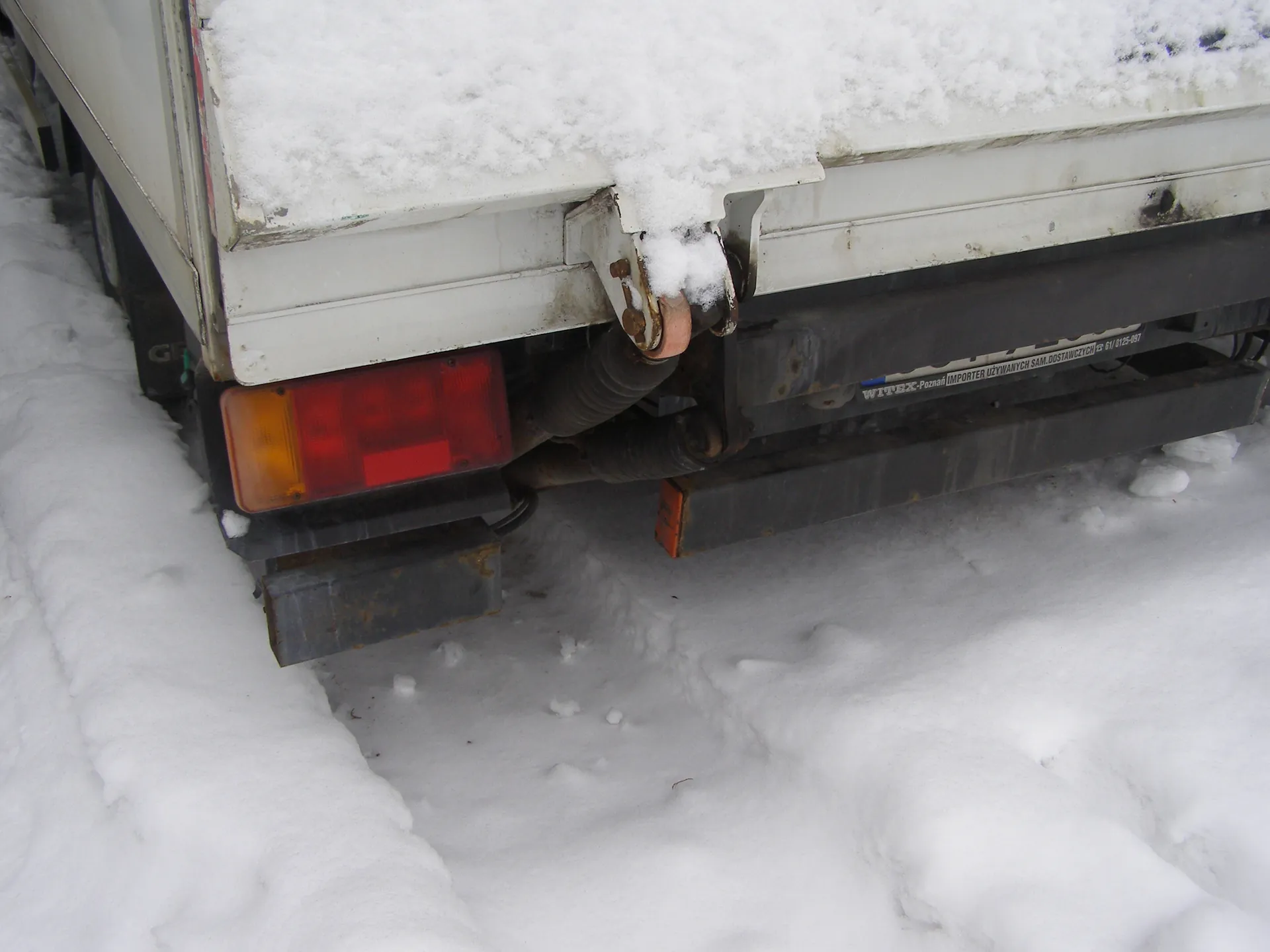 Tył białego pojazdu (przyczepa lub ciężarówka) pokryty śniegiem na krawędzi. Widoczne czerwone i pomarańczowe światła tylne. Czarny zderzak z rdzą i napisem "WITEX-Poznań IMPORTER UŻYWANYCH SAM. DOSTAWCZYCH 61/812-097". Śnieżna droga z odciskami opon i śladami zużycia.