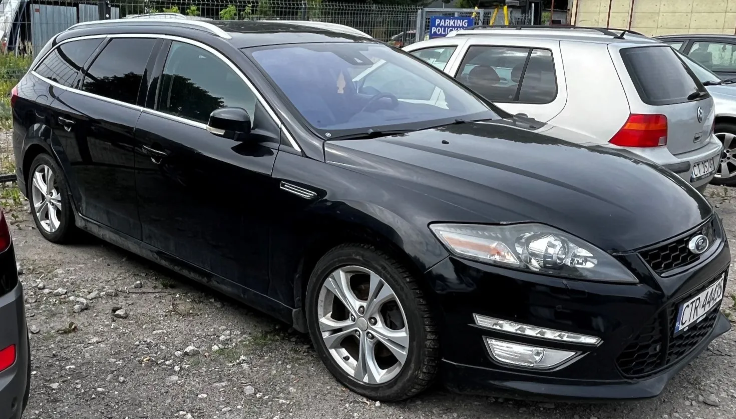 Czarny samochód osobowy w nadwoziu kombi marki Ford, zaparkowany na żwirowym placu. Auto jest widoczne z przodu i z boku. W tle znajduje się srebrny samochód oraz niebieska tablica z napisem 'PARKING POLICJI'.