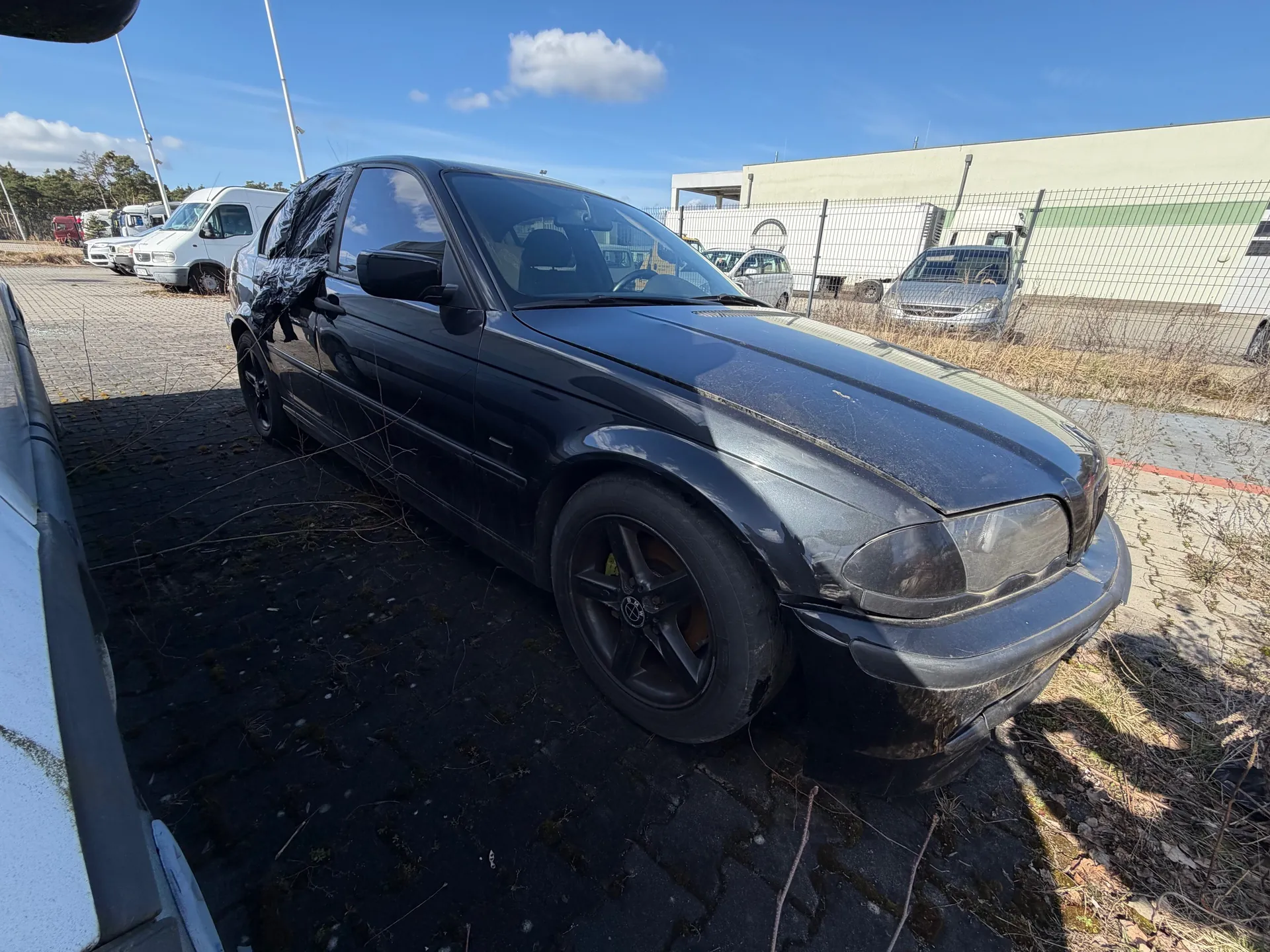 Czarny samochód osobowy z uszkodzonym przodem i tylnym oknem zakrytym czarną folią, zaparkowany na brukowanej powierzchni. W tle widoczne są inne pojazdy, biały van, metalowe ogrodzenie, budynek przemysłowy z zieloną pasem oraz niebo z kilkoma chmurami. Na ziemi sucha trawa i gałęzie.