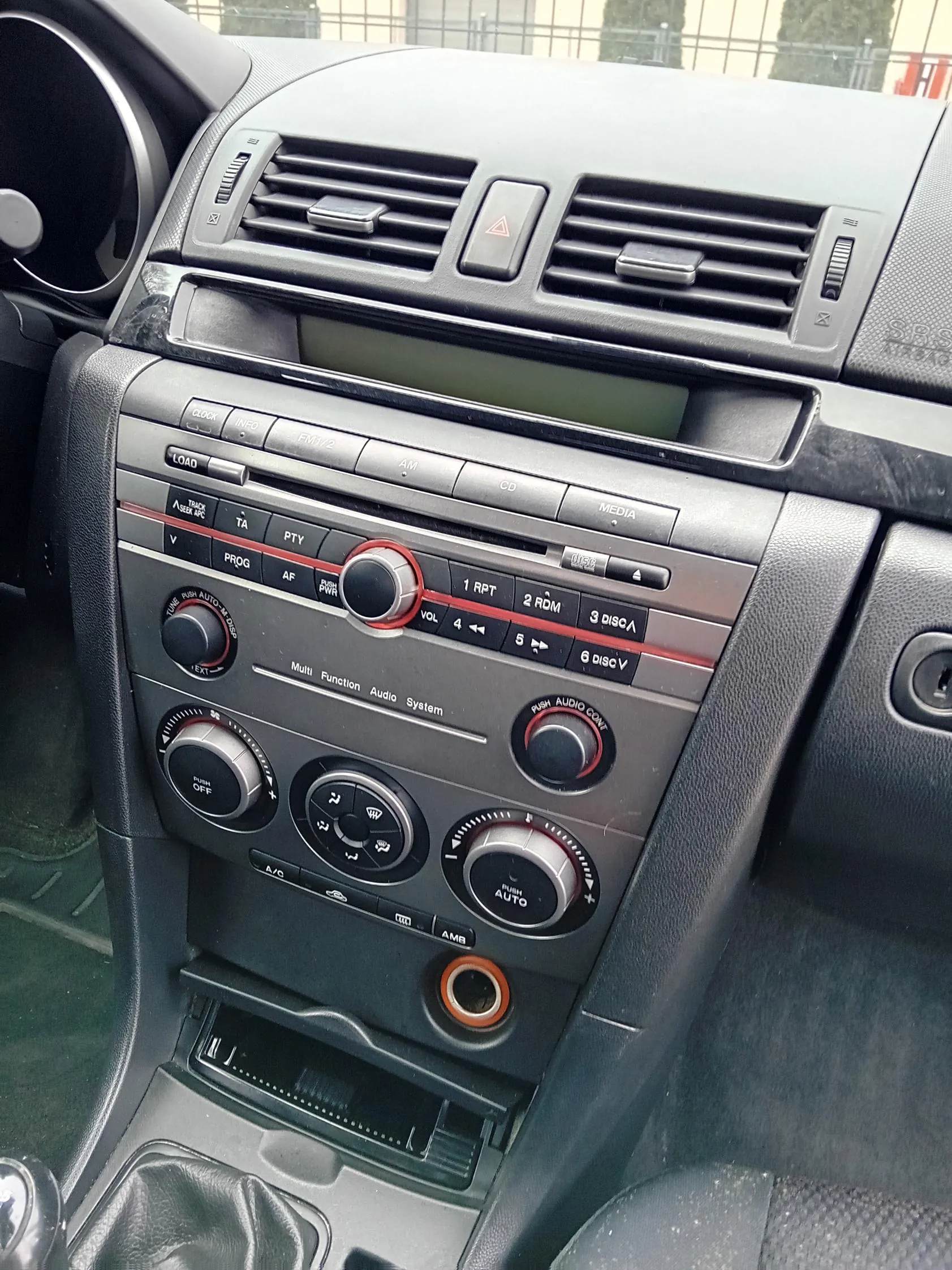 Wnętrze samochodu z widoczną deską rozdzielczą. Centralny panel zawiera system audio z przyciskami: LOAD, CLOCK, INFO, FM/AM, CD, MEDIA, 1 RPT, 2 RDM, 3 DISC, 4, 5, 6 DISC. Obok znajdują się pokrętła z napisami: TUNE PUSH AUTO, VOL, PUSH AUDIO CONT, A/C, AUTO. W górnej części panelu widoczne są kierunkowskazy, przycisk awaryjny (trójkąt), wentylatory i wyświetlacz. Na dole panelu znajduje się gniazdo z pomarańczowym obramowaniem oraz dźwignia zmiany biegów. W prawym górnym rogu widoczny napis SRS AIRBAG. W tle przez przednią szybę widać ogrodzenie i drzewa.