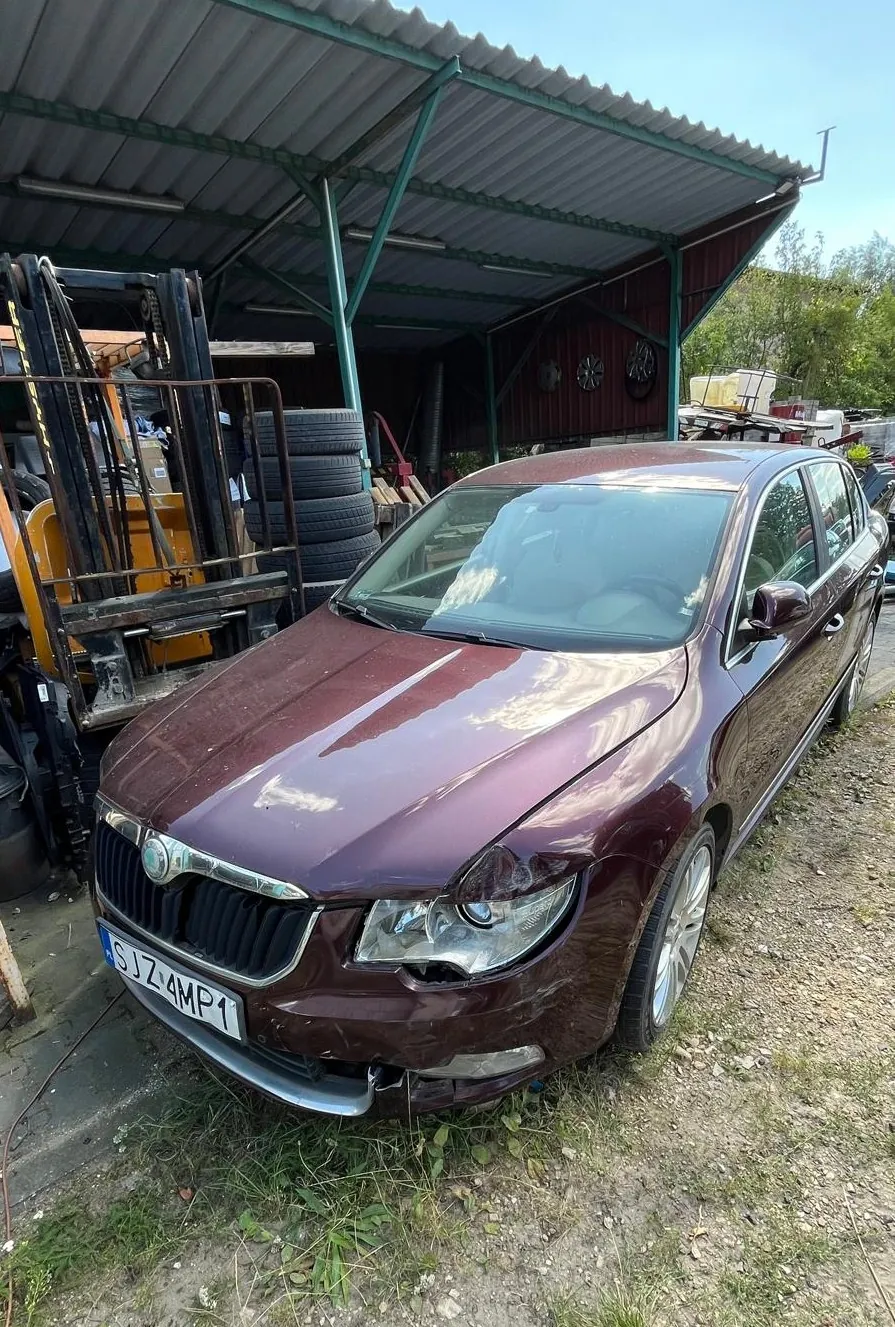 Bordowy samochód osobowy marki Skoda z uszkodzonym przodem, zaparkowany na nieutwardzonym terenie pod wiatą. Widoczne wgniecenie maski oraz pęknięty reflektor po prawej stronie. Samochód posiada tablicę rejestracyjną o numerze SJZ 4MP1. W tle po lewej stronie znajduje się wózek widłowy oraz stos opon.