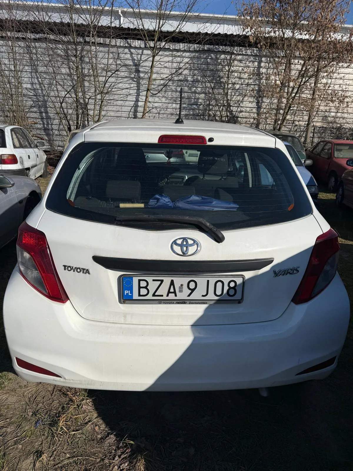 Biały samochód Toyota Yaris widziany od tyłu z tablicą rejestracyjną BZA 9J08 z kodem PL. W tle inne pojazdy, biały budynek z blachy falistej, bezlistne drzewa i jasne niebo. Na tablicy widoczny napis 'TOYOTA' po lewej stronie i 'YARIS' po prawej stronie. W środku tablicy rejestracyjnej znajduje się niebieski pas z literą 'PL' oraz logo Unii Europejskiej.