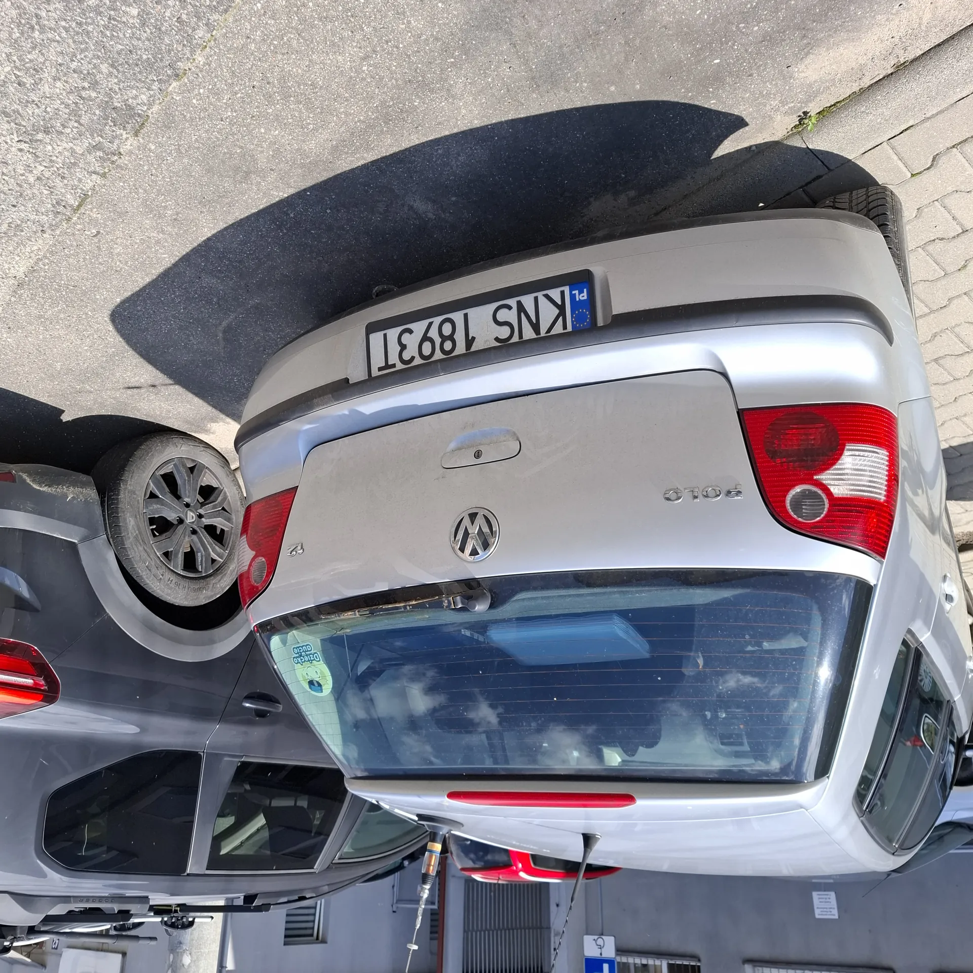 Srebrny samochód Volkswagen Polo z tablicą rejestracyjną KNS 1893T (PL) zaparkowany na ulicy. Obok niego stoi szary samochód. W tle widoczne są budynki i fragment drogi. Na tylnej szybie srebrnego samochodu znajduje się naklejka.