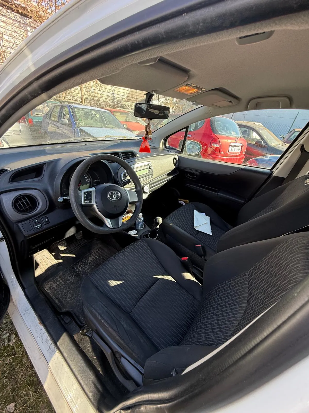 Wnętrze samochodu marki Toyota z widocznym kierownicą z logo, czarnymi siedzeniami w paski, deska rozdzielcza z przyciskami i wyświetlaczem. W tylnej części widoczne są inne zaparkowane samochody, w tym czerwony z tablicą rejestracyjną. Na siedzeniu pasażera leży biały papier, a na desce rozdzielczej wiszą czerwone ozdoby. Zewnętrzne tło obejmuje drzewa i ogrodzenie.