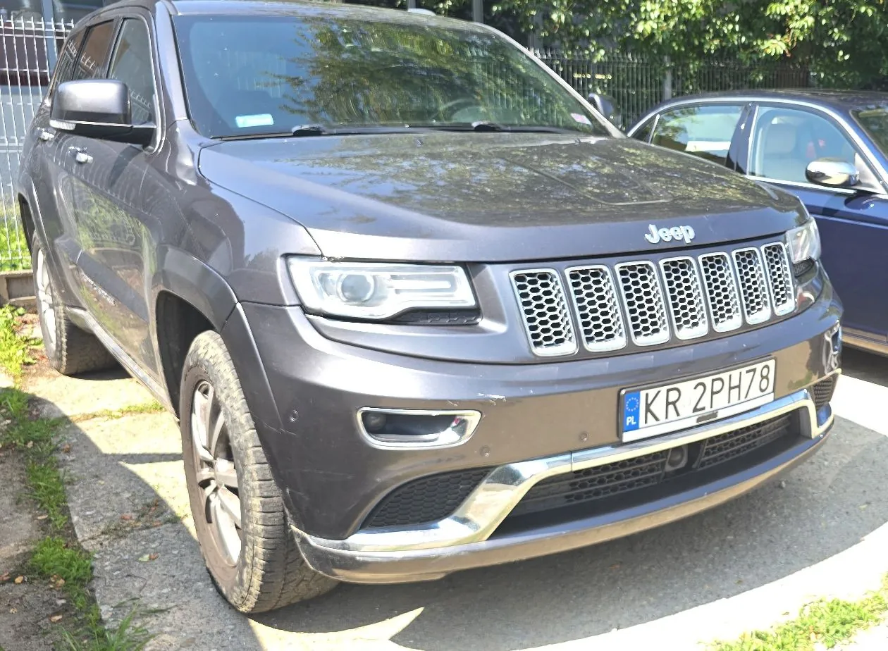 Srebrnoszary samochód marki Jeep Grand Cherokee zaparkowany na podjeździe. Pojazd ma polską tablicę rejestracyjną o numerze KR 2PH78. W tle widoczny jest metalowy płot oraz zielone drzewa.