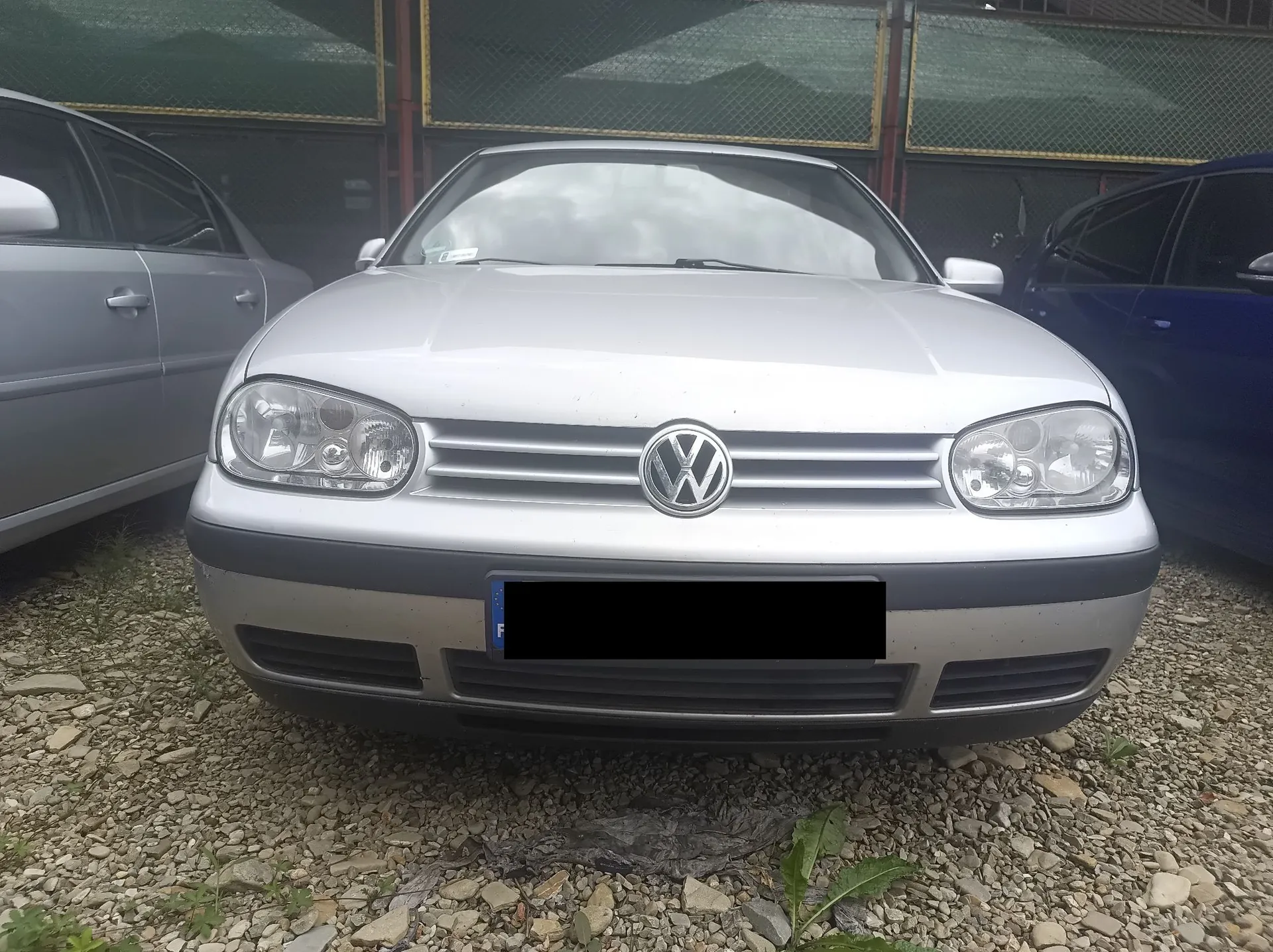 Srebrny samochód marki Volkswagen Golf widziany od przodu, zaparkowany na żwirowym podłożu. Po lewej stronie widoczny jest fragment innego srebrnego auta, a po prawej fragment niebieskiego samochodu. W tle znajduje się ogrodzenie z siatki. Tablica rejestracyjna pojazdu jest zakryta czarnym prostokątem.