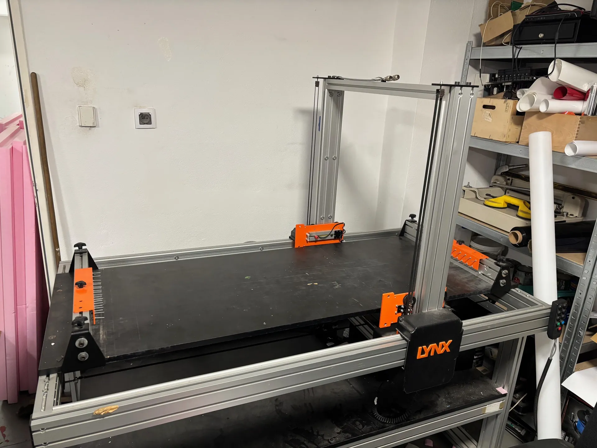 Przemysłowa maszyna z metalową ramą i pomarańczowymi elementami, z czarną powierzchnią roboczą. Na maszynie widoczny napis 'LYNX' pomarańczowymi literami. W tle biała ściana z gniazdem elektrycznym i przyciskiem, po lewej stronie różowe płyty izolacyjne, po prawej metalowa półka z kartonami, białymi rurami PVC, narzędziami i żółtym obiektem. Podłoga z czarnym materiałem.