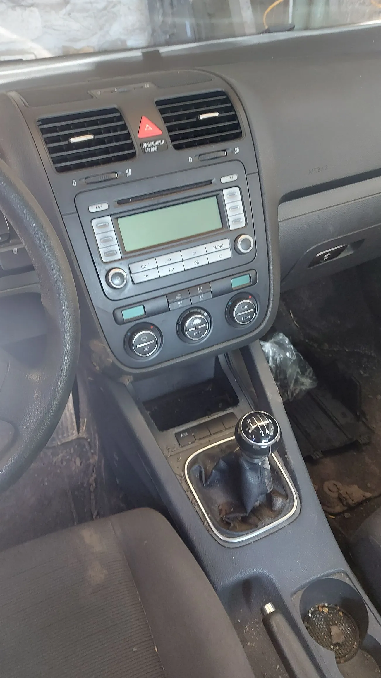 Wnętrze samochodu z widokiem na deskę rozdzielczą, konsolę środkową i lewark zmiany biegów. Na konsoli znajduje się radio z wyświetlaczem LCD, przyciski sterujące oraz pokrętła klimatyzacji. Poniżej znajduje się dźwignia zmiany biegów w manualnej skrzyni z czarną gałką. Wnętrze jest zakurzone, a obok konsoli w podłodze leży przezroczysta foliowa torba.