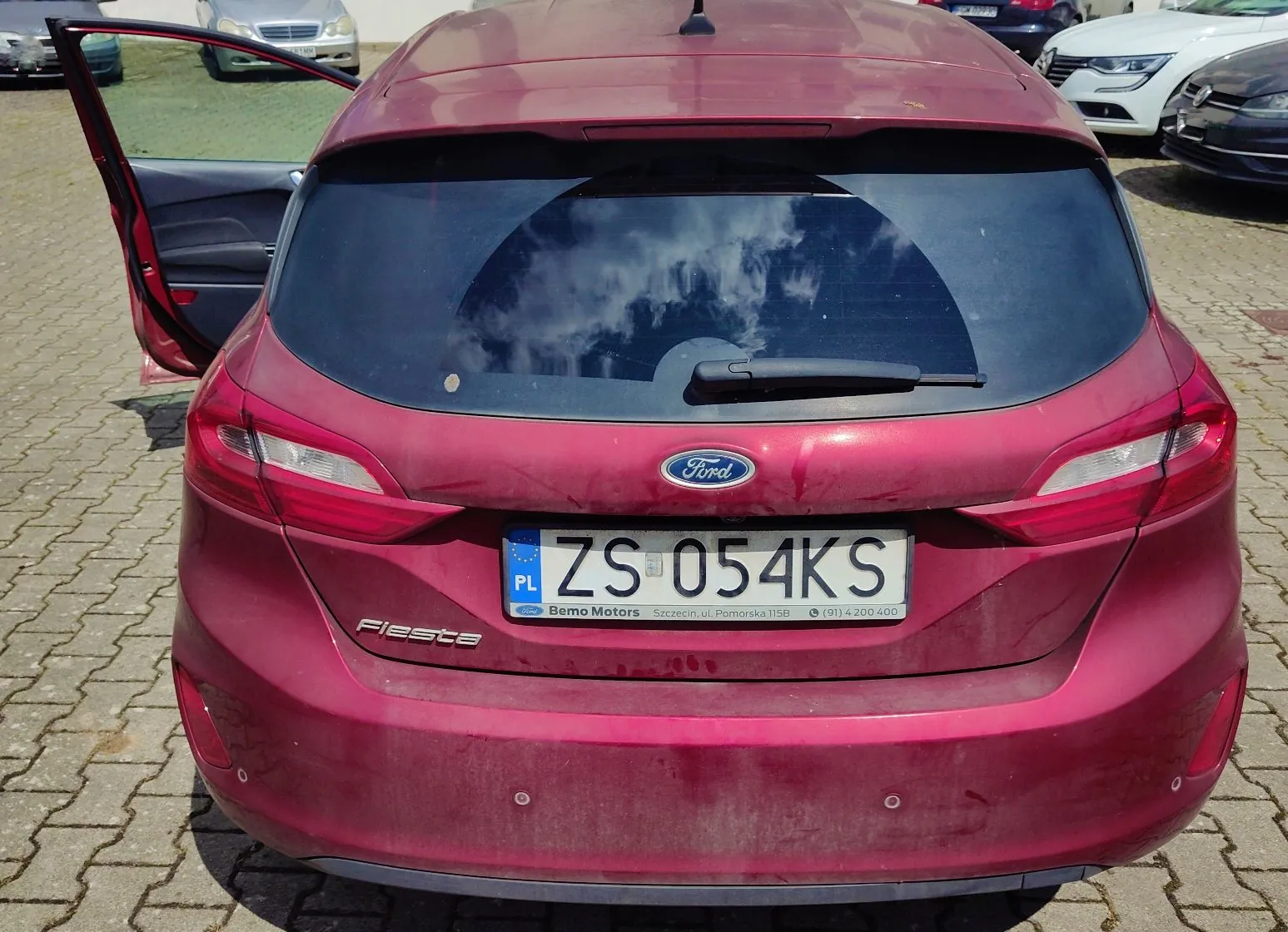Tylny widok czerwonego samochodu marki Ford Fiesta zaparkowanego na kostce brukowej. Samochód ma otwarte lewe tylne drzwi. Na klapie bagażnika znajduje się logo Ford oraz tablica rejestracyjna o numerze ZS 054KS z oznaczeniem PL. Poniżej tablicy widnieje napis: 'Bemo Motors Szczecin, ul. Pomorska 115B (91) 4 200 400'. W tle widoczne są fragmenty innych samochodów.