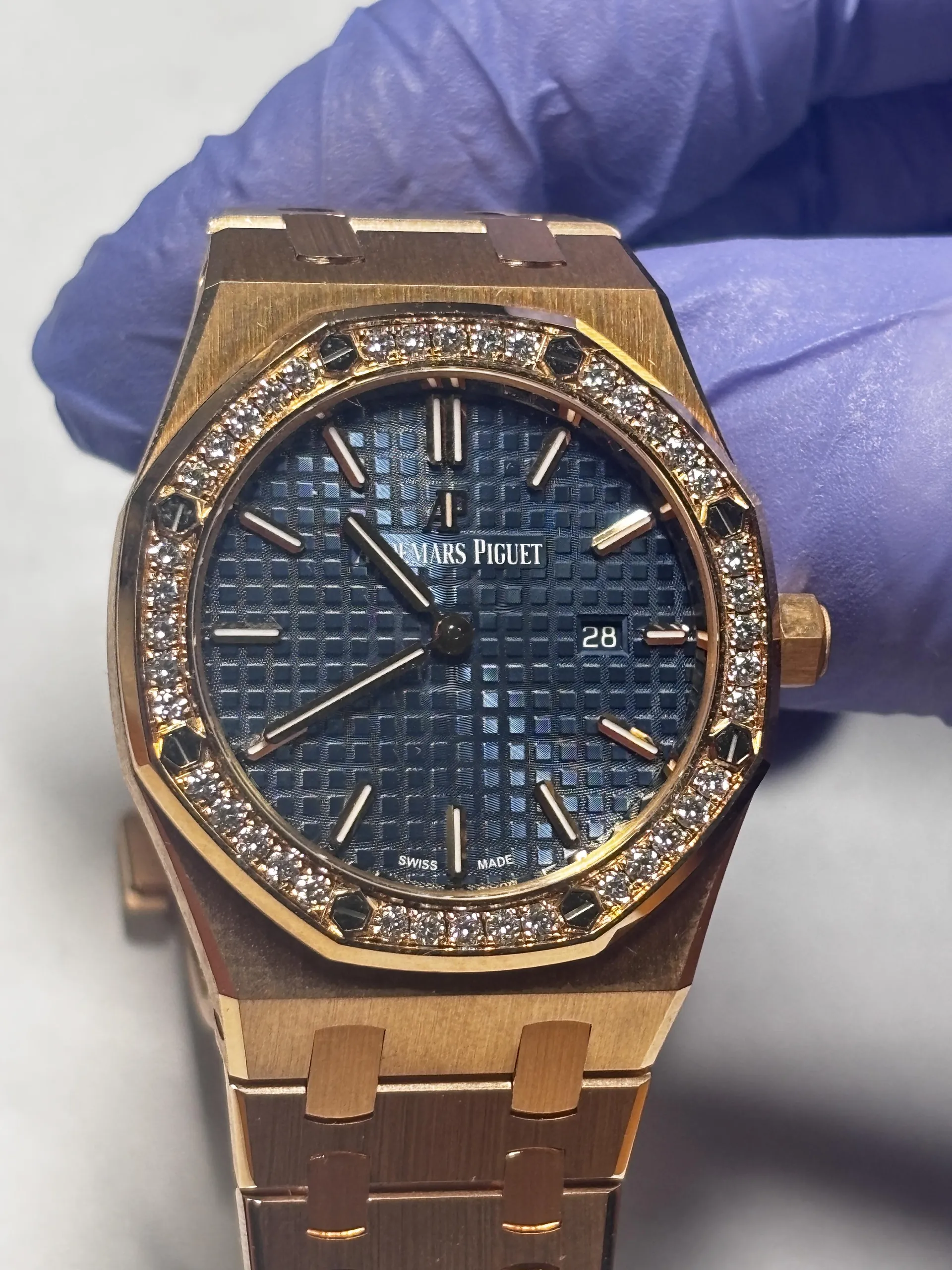 Złoty zegarek marki Audemars Piguet z niebieską tarczą w kratkę, ozdobiony diamentami na brzegu. Tarcza zawiera napisy 'Audemars Piguet', 'Swiss Made' oraz okienko daty z cyfrą '28'. Wskazówki i indeksy godzinowe są metalowe. Zegarek trzymana jest w ręce w niebieskiej rękawicy, na jasnoszarym tle.