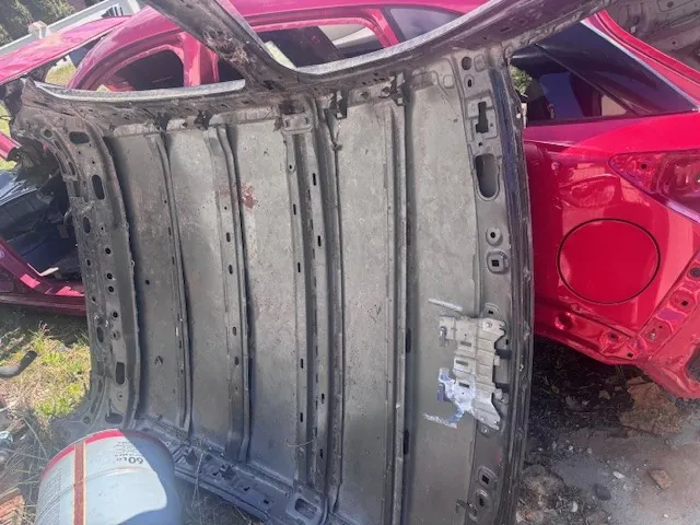 Zdjęcie przedstawia metalową konstrukcję samochodową, prawdopodobnie fragment drzwi lub nadwozia, z widocznymi otworami i śladami korozji. W tle znajduje się uszkodzony czerwony samochód z otwartymi drzwiami. Na dole zdjęcia widoczna jest opona z częściowo czytelnym napisem '60'. Tło stanowi ziemista powierzchnia z fragmentami trawy.
