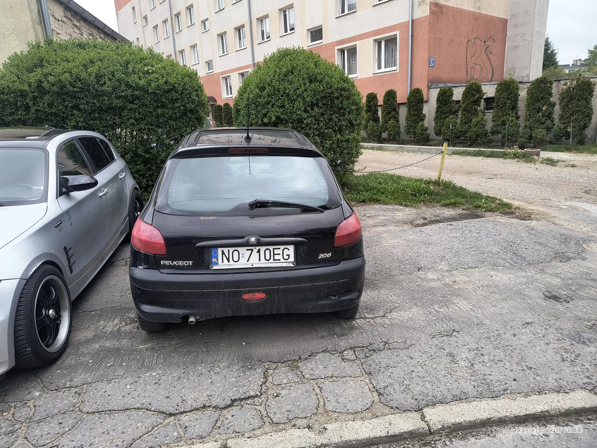 Zdjęcie przedstawia czarny samochód Peugeot 206 z tablicą rejestracyjną NO 710EG zaparkowany na pękniętym asfalcie. Po lewej stronie widoczny jest srebrny samochód. W tle budynek z czerwonymi i beżowymi elewacjami, oknami oraz zielonymi krzewami. Na prawo od budynku rosną wysokie krzewy, a na ziemi widoczna jest żwirek. W prawym dolnym rogu znajduje się znacznik czasu: 13 maj 2026, 10:33.