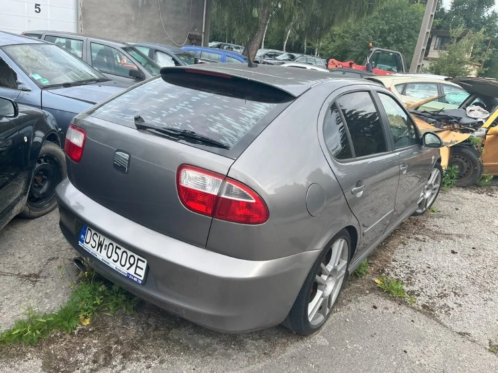 Srebrny samochód marki Seat Leon zaparkowany na betonowym placu wśród innych pojazdów. Widok z tyłu i z boku auta. Samochód posiada polską tablicę rejestracyjną o numerze DSW 0509E. W tle widoczne są inne samochody, w tym uszkodzony żółty pojazd oraz fragmenty budynków i drzewa.