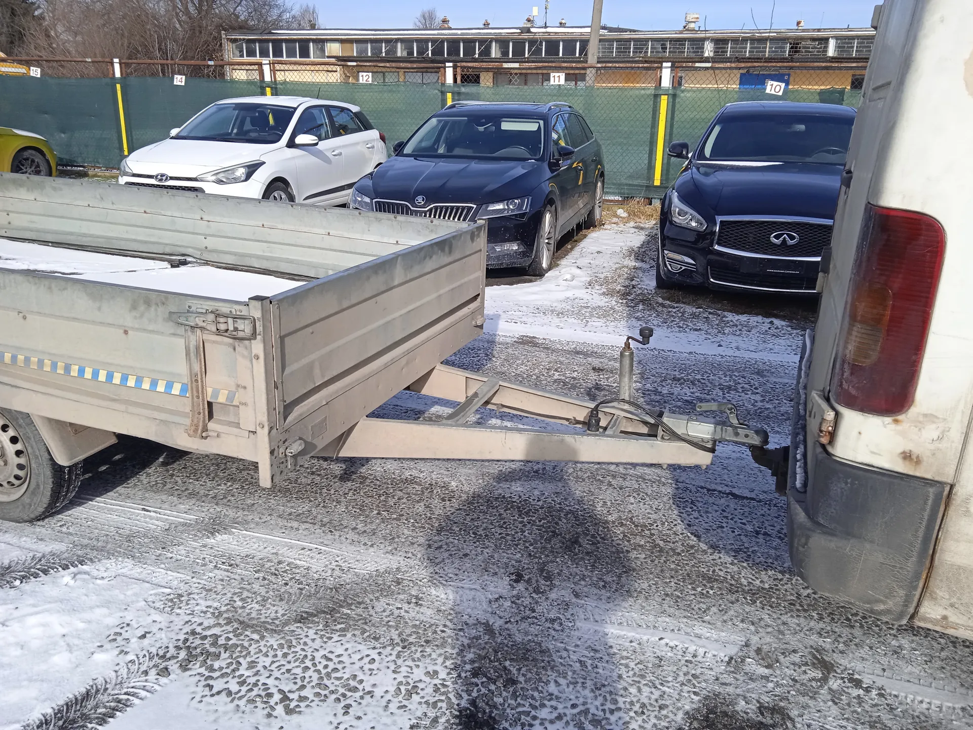 Stojące na zimowym parkingu pojazdy: metalowa przyczepa z niebiesko-żółtymi paskami przyczepiona do białego furgonu po prawej stronie, biały samochód Hyundai, ciemny samochód Skoda i ciemny samochód Infiniti. Częściowo widoczny żółty samochód po lewej stronie. Na tle zielone ogrodzenie z białymi numerami 10, 11, 12, 14, 15 oraz budynki. Na ziemi śnieg i lód.