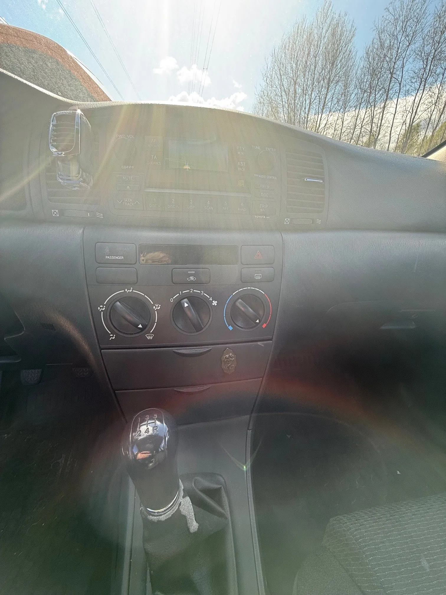 Wnętrze samochodu Toyota z widokiem na deskę rozdzielczą i dźwignię biegów. Deska rozdzielcza zawiera trzy okrągłe pokrętła klimatyzacji, radio z logo "TOYOTA" i przyciskami, oraz wskaźnik "PASSENGER" dla poduszki powietrznej pasażera. Dźwignia biegów manualnych oznaczona cyframi 1-5 i literą R, z ozdobnym pokryciem. Przez przednią szybę widoczne są drzewa bez liści, niebo z chmurami i linie energetyczne. Słoneczne światło powoduje pojawienie się poślizgów na obiektywie.